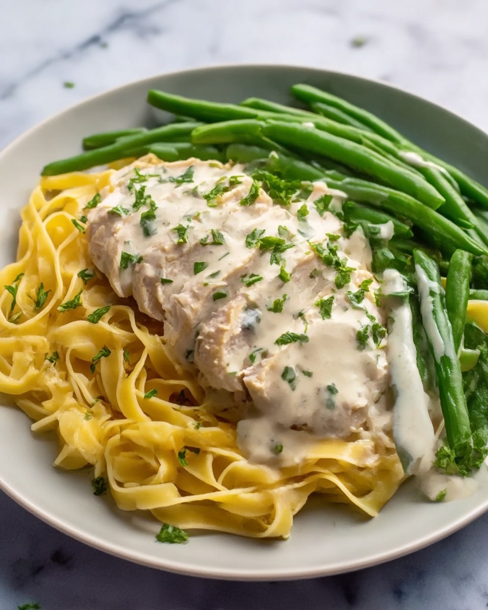 The image shows a white plate with four layers of food on a white marbled surface. The bottom layer is a bed of yellow fettuccine pasta with a smooth texture. On top of the pasta is a piece of light brown chicken, sliced into thin strips and covered with a creamy white sauce that has small green herb pieces sprinkled on it. Next to the chicken and pasta are bright green cooked green beans with a similar creamy white sauce drizzled over them. The whole dish is garnished with finely chopped fresh green herbs scattered over the top. Photo taken with an iphone --ar 4:5 --v 7
