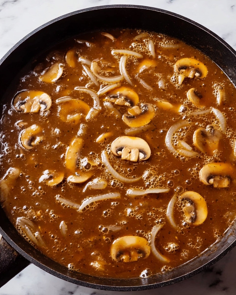The image shows a black pan filled with a rich brown sauce containing slices of light brown mushrooms and thin strips of translucent white onions. The sauce looks thick with small bubbles on the surface and a glossy texture that reflects light. The mushrooms are scattered throughout the sauce, with some pieces overlapping the onion slices. The pan sits on a white marbled surface. photo taken with an iphone --ar 4:5 --v 7