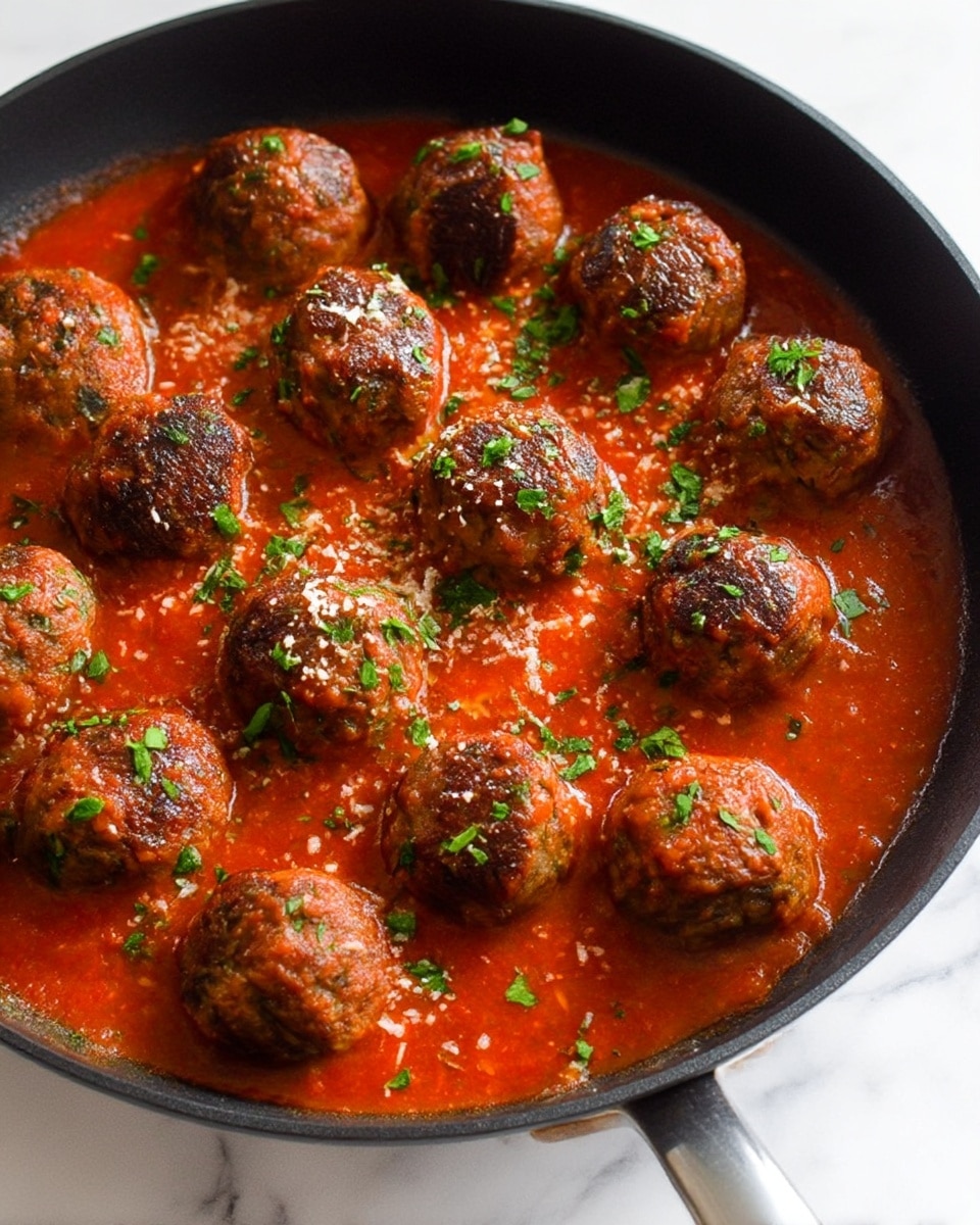 A black skillet filled with about thirteen browned meatballs sitting in a thick bright red tomato sauce. The meatballs have a caramelized crust on top with some small green herbs scattered across them and the sauce. The sauce looks smooth with a slightly chunky texture. Small bits of grated white cheese are sprinkled lightly over the meatballs and sauce. The skillet handle is visible at the bottom right, and the skillet rests on a white marbled surface. photo taken with an iphone --ar 4:5 --v 7