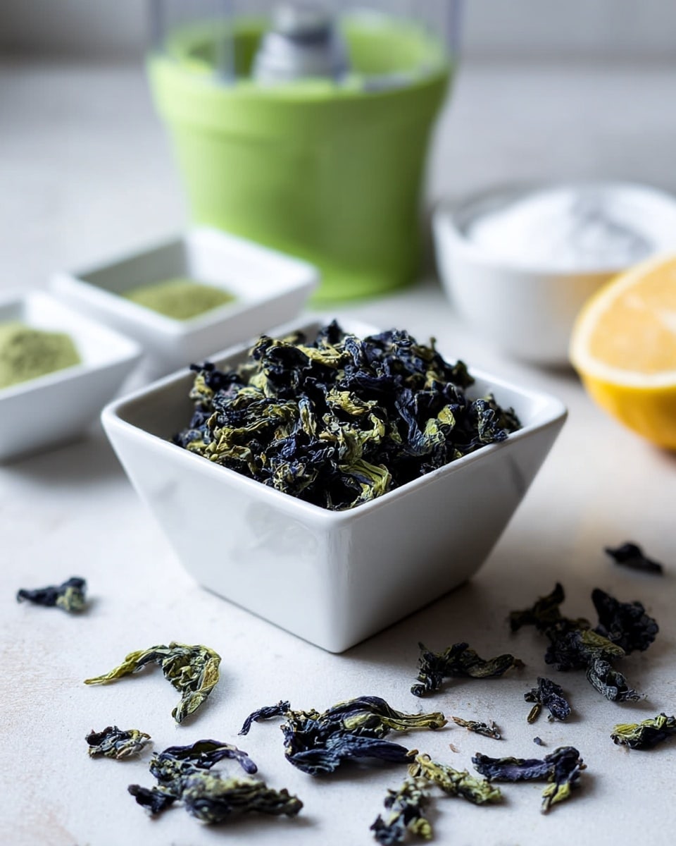 A white square bowl is filled with dried butterfly pea flowers showing dark blue and light green colors with a rough, shriveled texture. Some dried flowers spill around the bowl on a white marbled surface. In the blurred background, there is a green juicer bowl along with two small white square bowls, one holding green powder and the other containing a white granular substance. A yellow lemon, partly visible on the right, adds a bright touch. The photo has soft natural light and a clean, calm setting. photo taken with an iphone --ar 4:5 --v 7