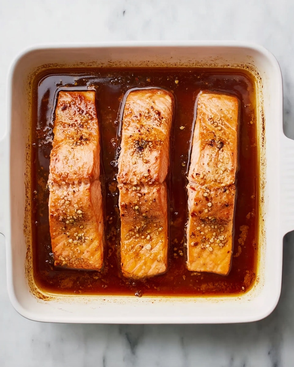 The image shows a white rectangular baking dish with four pieces of cooked salmon arranged in a single layer, side by side, each piece with a light orange-pink color and slightly browned edges. The salmon sits in a dark reddish-brown sauce that fills the bottom of the dish, with a slightly oily texture visible. The baking dish is placed on a white marbled surface. The photo taken with an iphone --ar 4:5 --v 7