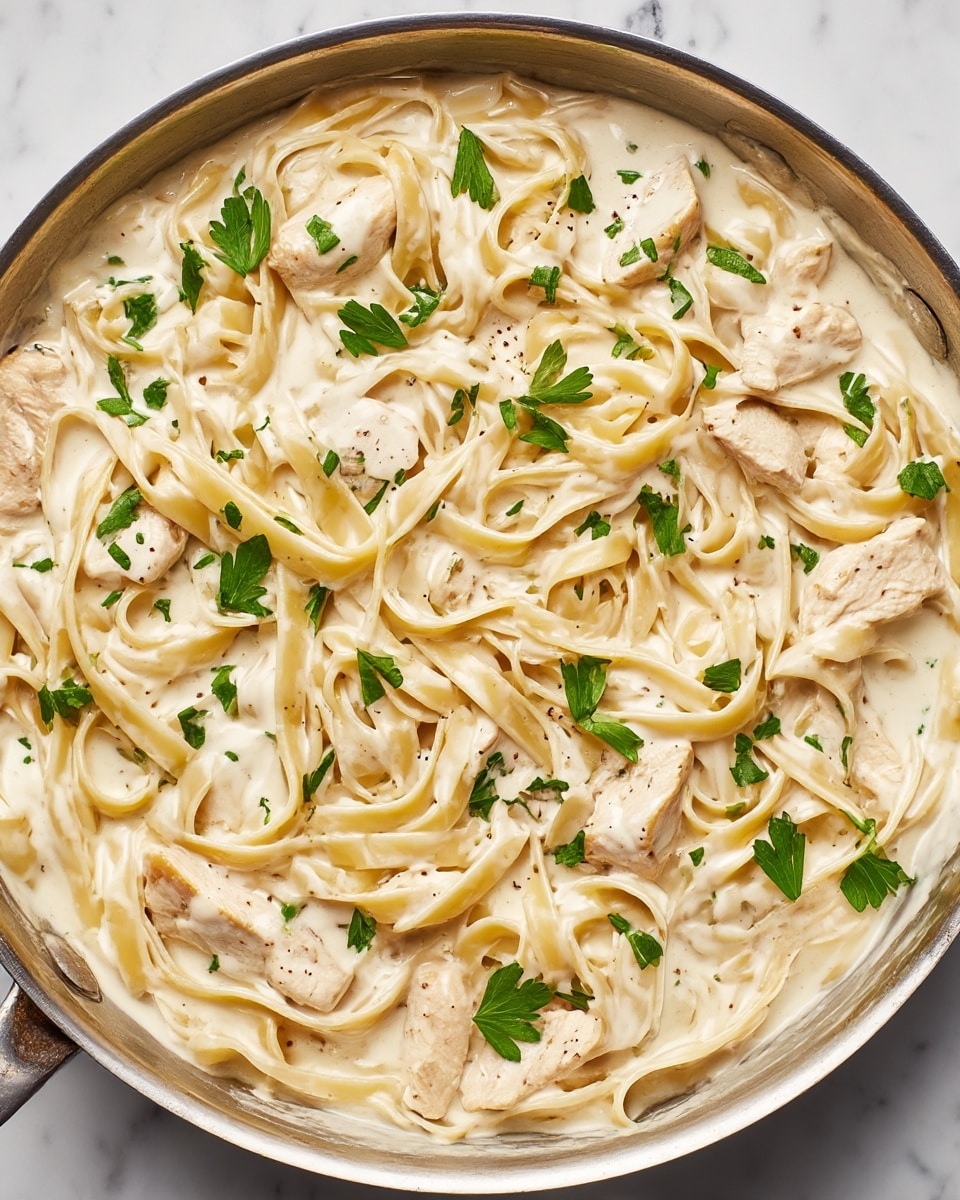 This image shows a pan filled with creamy fettuccine pasta and pieces of cooked chicken. The pasta is long and flat, coated in a thick white cream sauce with specks of black pepper. Chicken pieces are light brown on the edges and spread throughout the dish evenly. Fresh green parsley leaves are sprinkled on top, adding a pop of color. The pan has a metal rim and the dish sits on a white marbled surface. Photo taken with an iphone --ar 4:5 --v 7