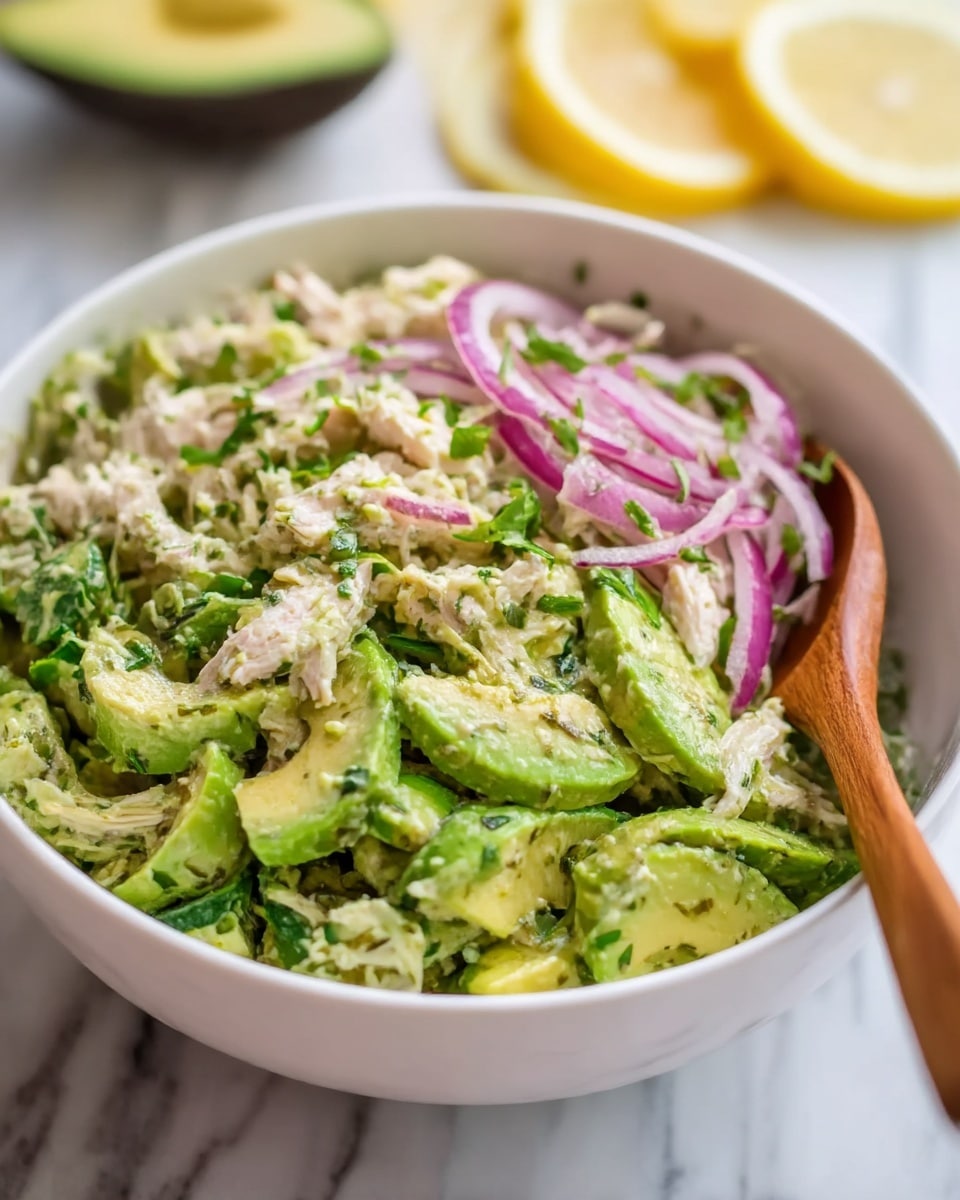 A white bowl filled with a fresh avocado salad, showing about three main layers. The bottom layer is light green slices of avocado mixed with herbs, the middle layer has shredded light pink chicken pieces, and the top layer contains thin slices of purple onion and small green herbs scattered across. A wooden spoon rests inside the bowl on the right side. The background is a white marbled surface with blurred slices of lemon at the back. photo taken with an iphone --ar 4:5 --v 7