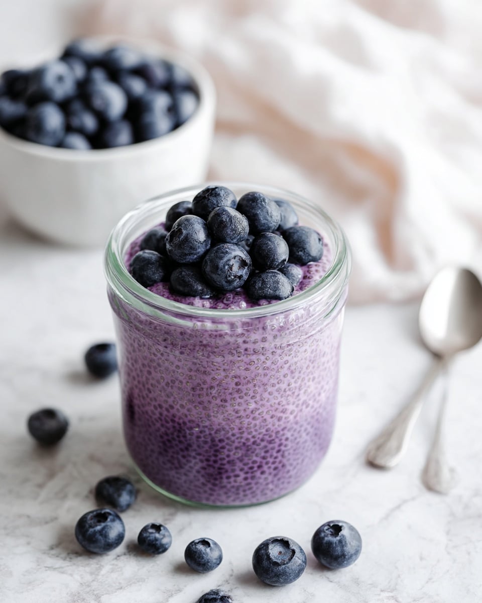 A clear glass jar filled with two layers of purple chia pudding topped with a cluster of fresh, plump blueberries. The pudding has a thick, textured look with tiny chia seeds spread evenly throughout. Around the jar are loose blueberries resting on a white marbled surface. In the background, a small white bowl filled to the top with blueberries and a blurred silver spoon with a soft white cloth are visible. The overall image is light and bright with a soft focus on the background. photo taken with an iphone --ar 4:5 --v 7