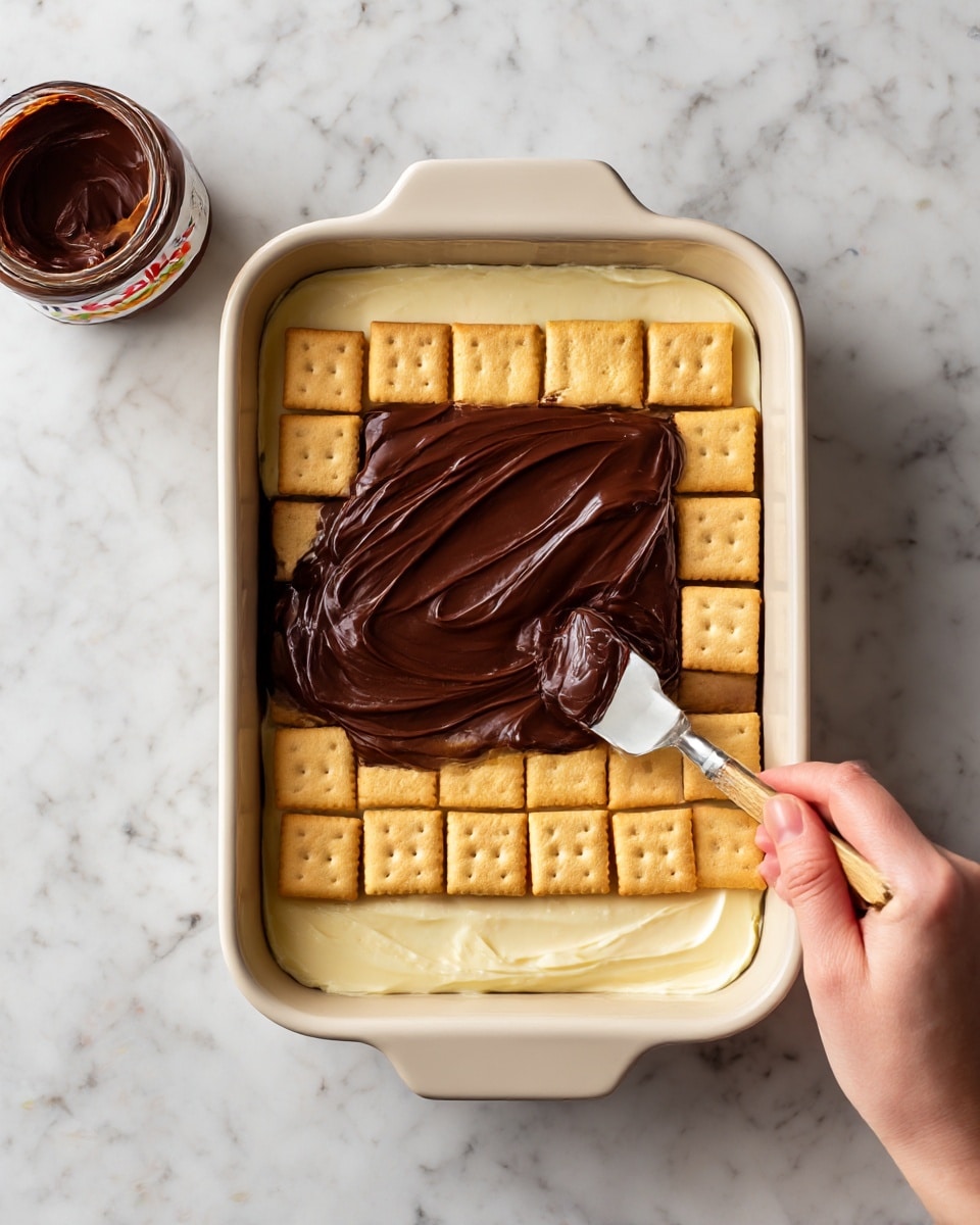 A white rectangular baking dish with a layer of light yellow custard at the bottom, topped with square beige graham crackers neatly arranged side by side. A woman's hand is spreading a thick, smooth, dark brown chocolate layer over the graham crackers with a flat spatula. The background is a white marbled surface, and a jar of dark chocolate spread is visible at the top left corner. Photo taken with an iphone --ar 4:5 --v 7
