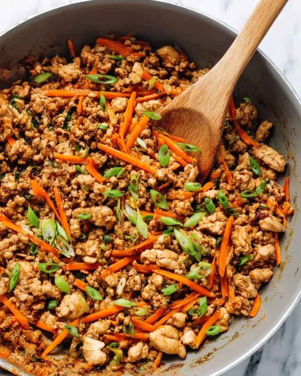 A gray pan holds a mix of cooked ground meat that is light brown with a slightly crumbly texture, thin strips of bright orange carrots, and sliced green onions scattered on top. A wooden spoon is resting in the pan, angled from the right side, showing pieces of the mixture on it. The background surface is a white marbled texture. Photo taken with an iphone --ar 4:5 --v 7