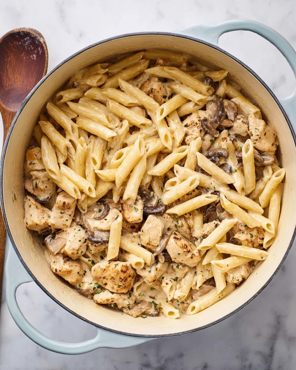 A large white pot filled with a pasta dish showing two main layers: the top is light yellow penne pasta mixed with creamy sauce, and underneath are thick pieces of chicken seasoned with herbs and mixed with dark brown bits of mushrooms. The creamy sauce lightly coats all ingredients, giving a smooth texture, and the pot sits on a white marbled surface. A wooden spoon with a dark handle rests next to the pot, adding a rustic touch. photo taken with an iphone --ar 4:5 --v 7