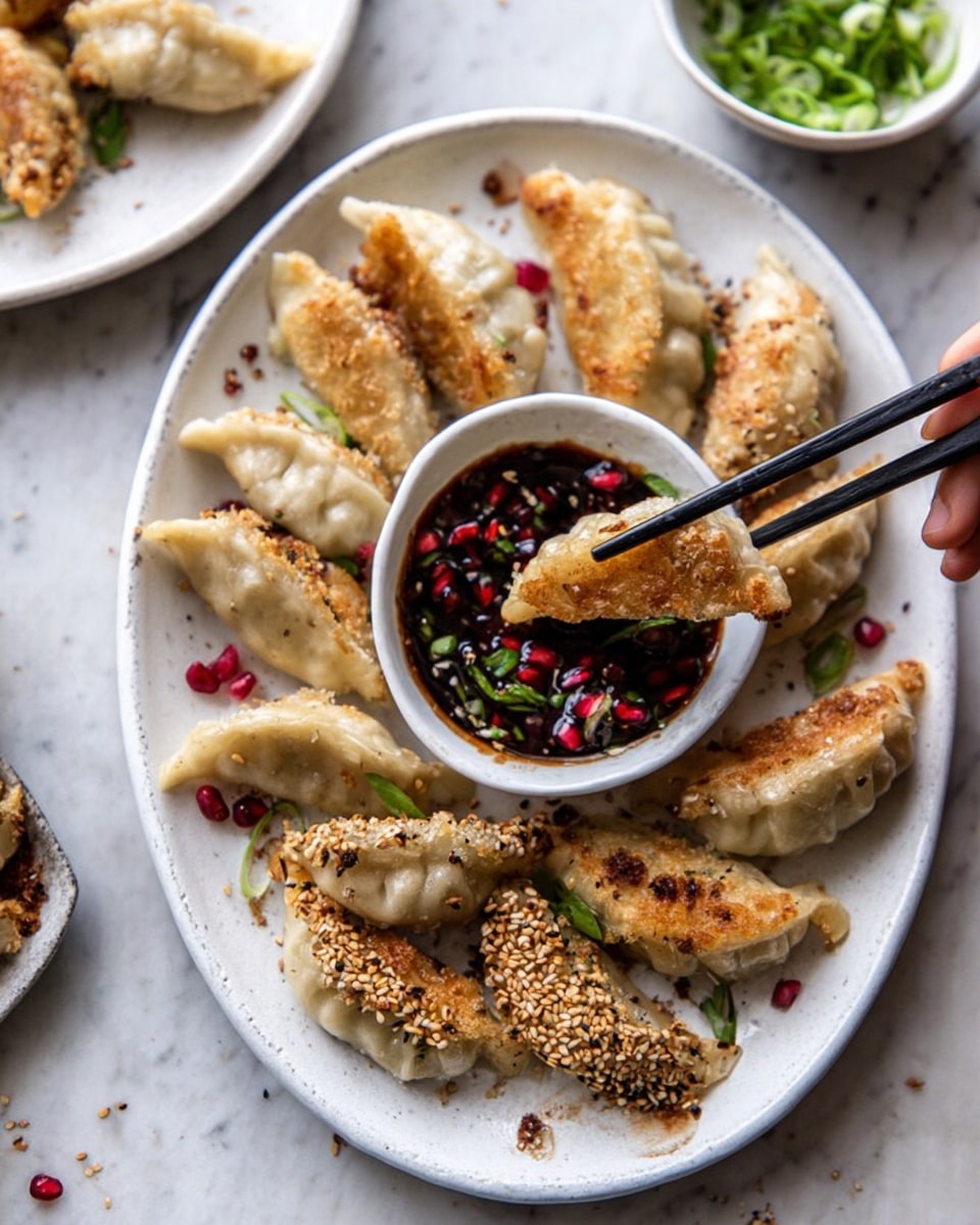 The image shows an oval white plate filled with golden brown dumplings, some of which are coated with sesame seeds on one side, arranged around a small white bowl of dark dipping sauce with red pomegranate seeds and green herbs inside. One dumpling is being held above the bowl by black chopsticks held by a woman's hand. The dumplings have a slightly crispy texture on the sesame side and a soft, smooth pale dough on the other side. The plate is on a white marbled surface, and there is another white plate partially visible with more dumplings in the background, along with a small white bowl of chopped green onions. photo taken with an iphone --ar 4:5 --v 7