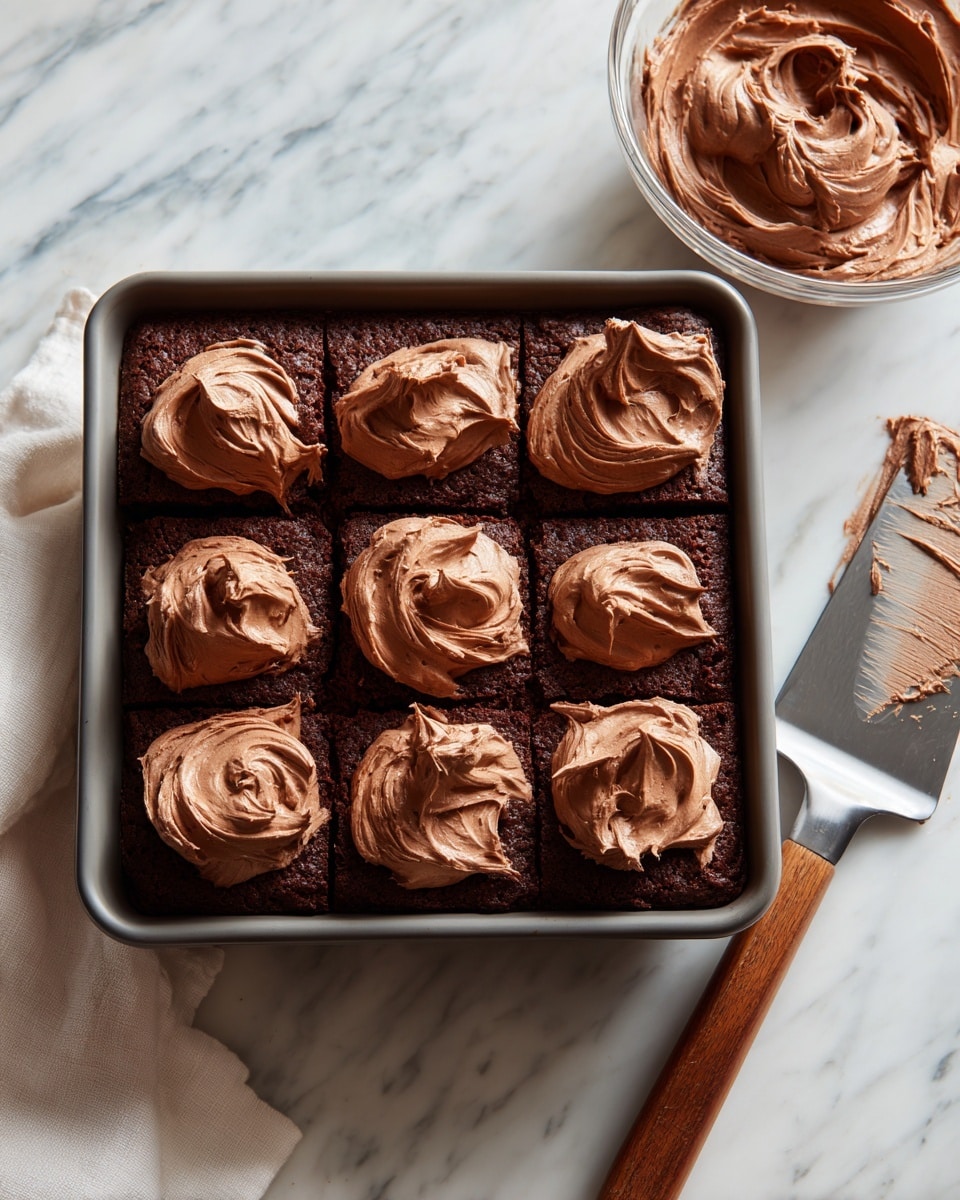 A square chocolate cake in a gray baking pan forms the base layer with a dark brown, slightly textured surface. On top, there are multiple thick and creamy swirls of light brown chocolate frosting spread unevenly across the cake’s surface in nine dollops. To the top right side of the pan, a clear glass bowl holds more of the same smooth chocolate frosting with visible swirls inside. A spatula with a gray blade and wooden handle rests to the right on the white marbled surface, some frosting spread lightly on its blade. A white cloth is partially visible at the bottom left corner. Photo taken with an iphone --ar 4:5 --v 7