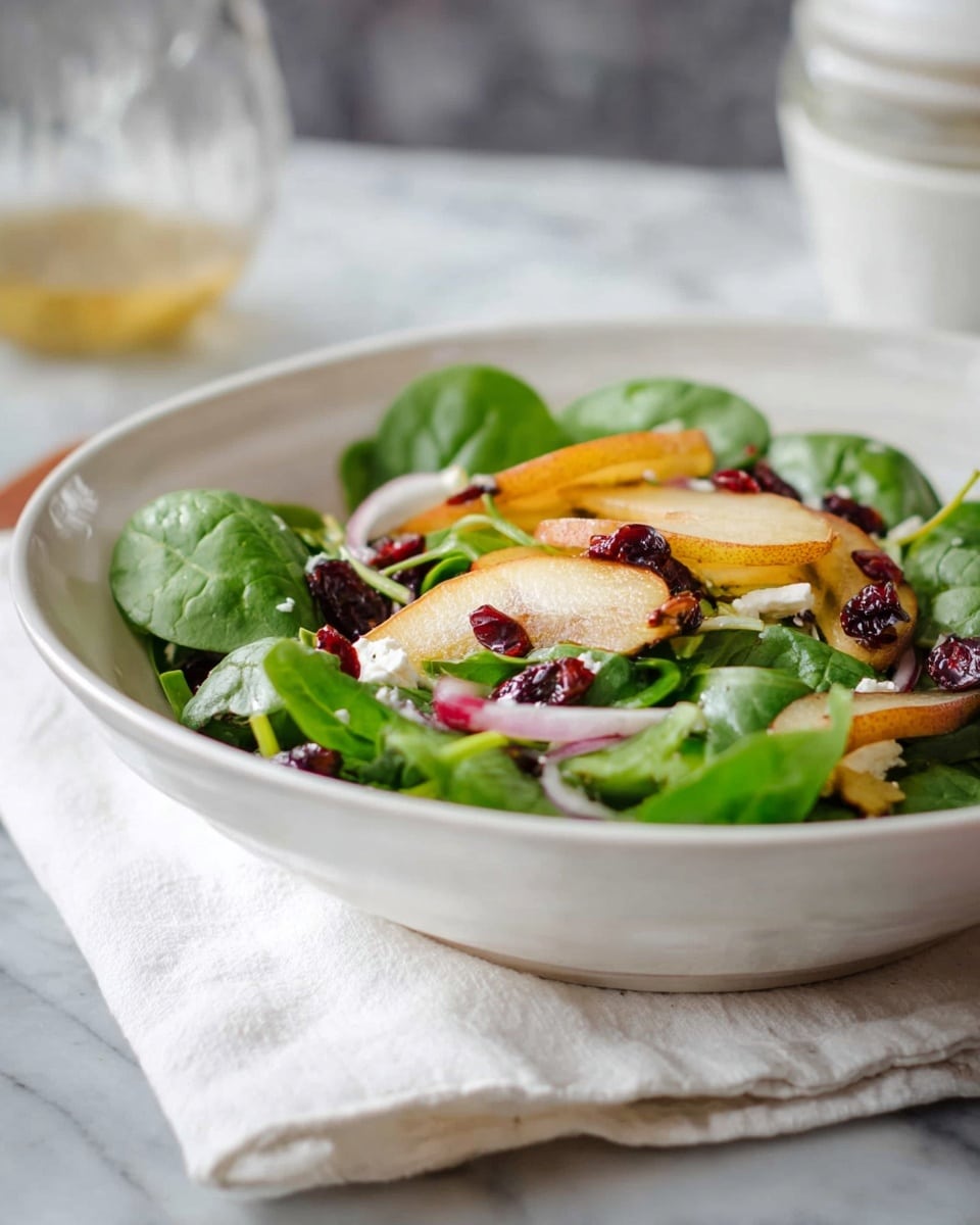 A white bowl holds a fresh salad with a base of bright green spinach leaves. On top, there are soft-looking slices of golden-brown roasted pear, scattered dried red cranberries, thin rings of light purple onion, and small chunks of white cheese. The salad sits on a white folded cloth, placed over a surface with a white marbled texture. In the background, there is a blurred clear glass container with dressing and a white bowl. Photo taken with an iphone --ar 4:5 --v 7