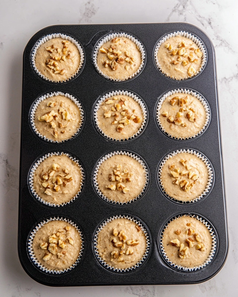A black metal muffin tray holds twelve white paper liners, each filled with light brown batter that looks thick and slightly lumpy. Every muffin is topped with a few small pieces of chopped walnuts that add texture and a rough pattern on top. The tray rests on a white marbled surface, enhancing the soft, natural colors of the batter and nuts. The muffin batter fills each liner almost to the top, showing that they are ready to bake. photo taken with an iphone --ar 4:5 --v 7
