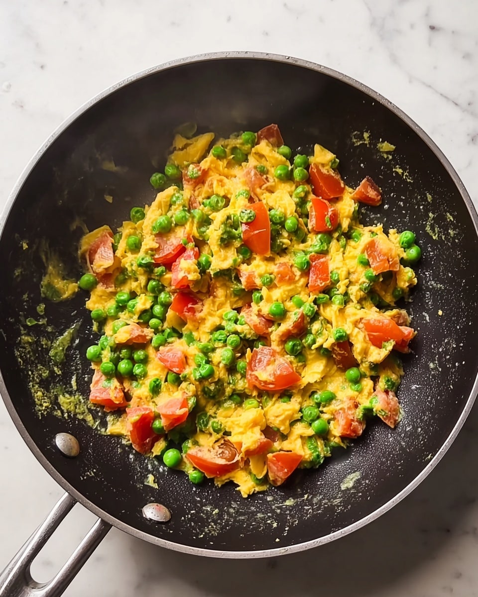 Inside a black frying pan with two silver handles, there is a mix of scrambled yellow eggs, bright green peas, and chunks of red tomatoes. The eggs are soft and mixed evenly with the vegetables. The peas are round and fresh, scattered throughout with the tomato pieces that have smooth, shiny skin. The pan sits on a white marbled surface, and there is slight steam rising from the warm food. photo taken with an iphone --ar 4:5 --v 7