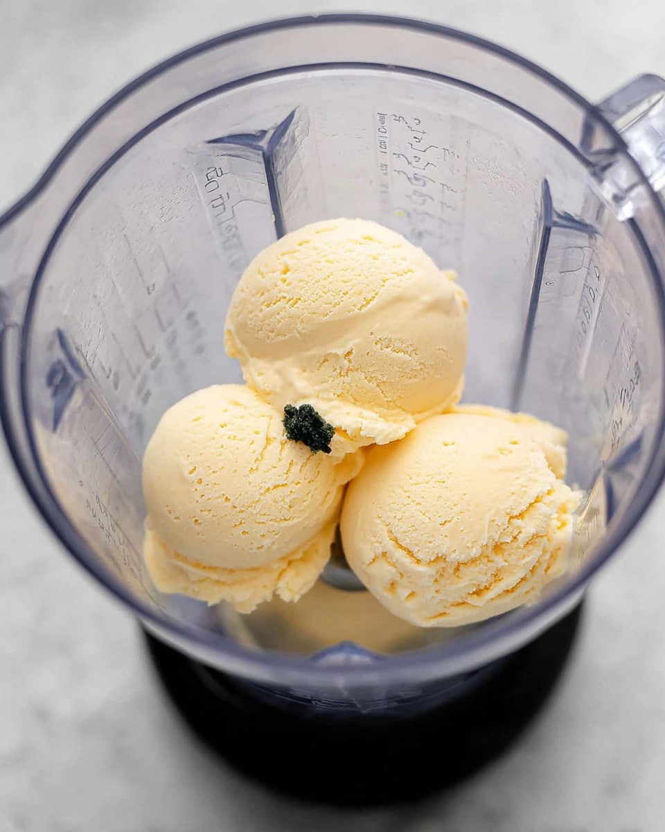 The image shows a clear blender container with three scoops of pale yellow ice cream placed inside. The texture of the ice cream is smooth with slight holes and soft ridges visible, and there is a small dollop of dark green paste sitting on top of one scoop near the center. The blender's measurement markings are faintly visible on the sides, and the background is a clean white marbled surface, adding contrast to the ice cream and the blender. photo taken with an iphone --ar 4:5 --v 7