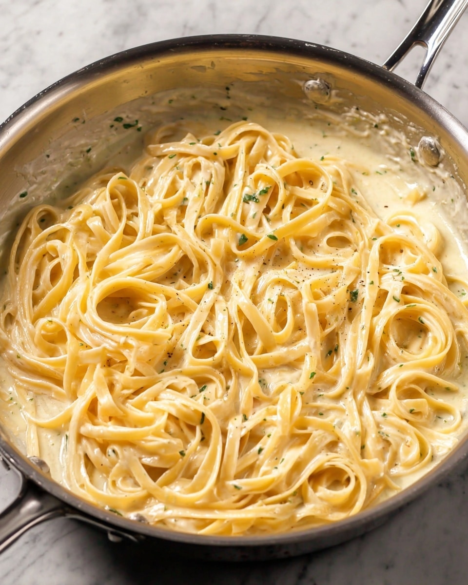 A shiny silver pan is filled with creamy light yellow pasta noodles mixed evenly with a smooth sauce. The sauce covers all the noodles, giving a shiny and thick look with small green herb bits and a few specks of black pepper scattered throughout. The noodles are long, flat, and slightly curled, lying close together in the pan. The pan sits on a white marbled surface. Photo taken with an iphone --ar 4:5 --v 7