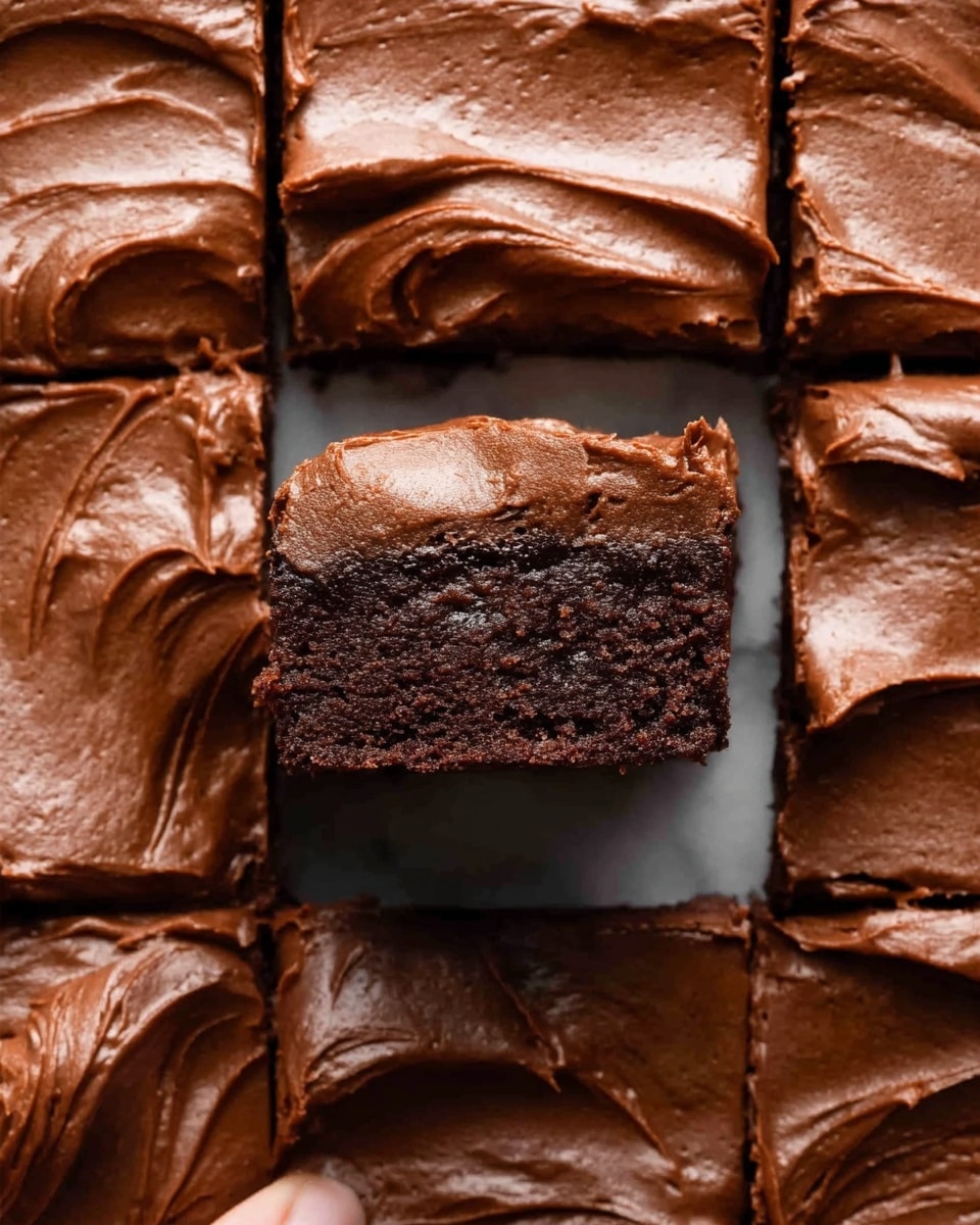 The image shows a close-up of a rich chocolate dessert with thick, smooth chocolate frosting spread in thick swirls all over the top surface. A single square piece is lifted slightly above the rest, revealing a soft, dense, and fudgy dark brown layer beneath the glossy chocolate icing. The creamy top layer has a slightly shiny texture with some peaks and valleys, showing its smooth and thick spread. The dessert is placed on a white marbled surface, and a woman's hand is holding the lifted piece gently. Photo taken with an iphone --ar 4:5 --v 7