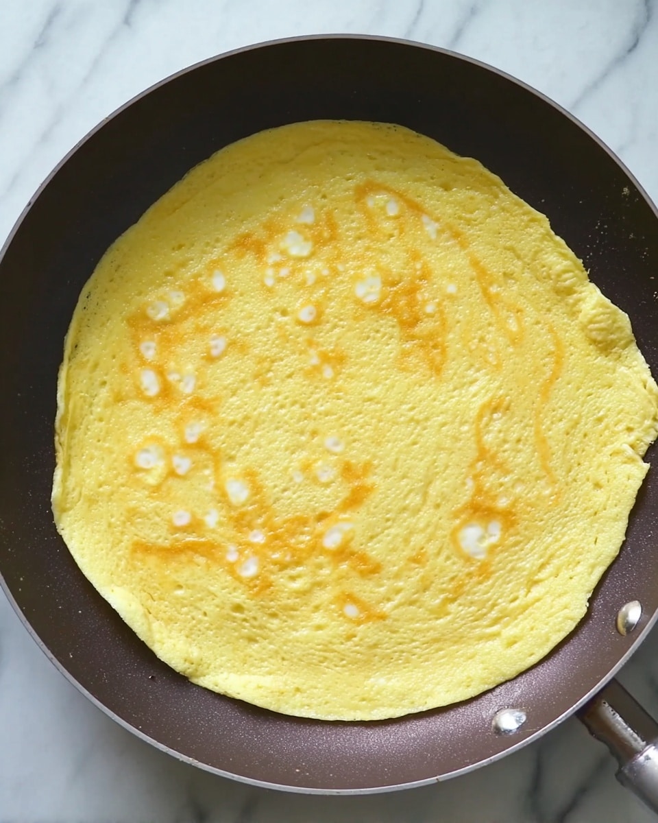 A large, thin omelette with a light yellow color and small white spots from cooked egg whites is inside a dark frying pan. The omelette spreads evenly across the pan's surface, with a slightly uneven edge on the right side. The frying pan sits on a white marbled surface. photo taken with an iphone --ar 4:5 --v 7