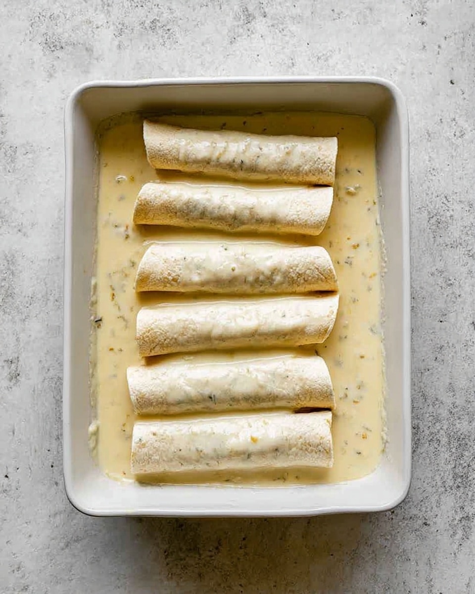 A white square baking dish holds eight rolled tortillas placed evenly in two rows on a creamy light beige sauce that looks thick and speckled with small bits. The tortillas are pale, smooth, and soaking in the sauce, some partially submerged, showing their soft texture. The dish sits on a white marbled surface, and the image is taken from above showing the arrangement clearly. Photo taken with an iphone --ar 4:5 --v 7