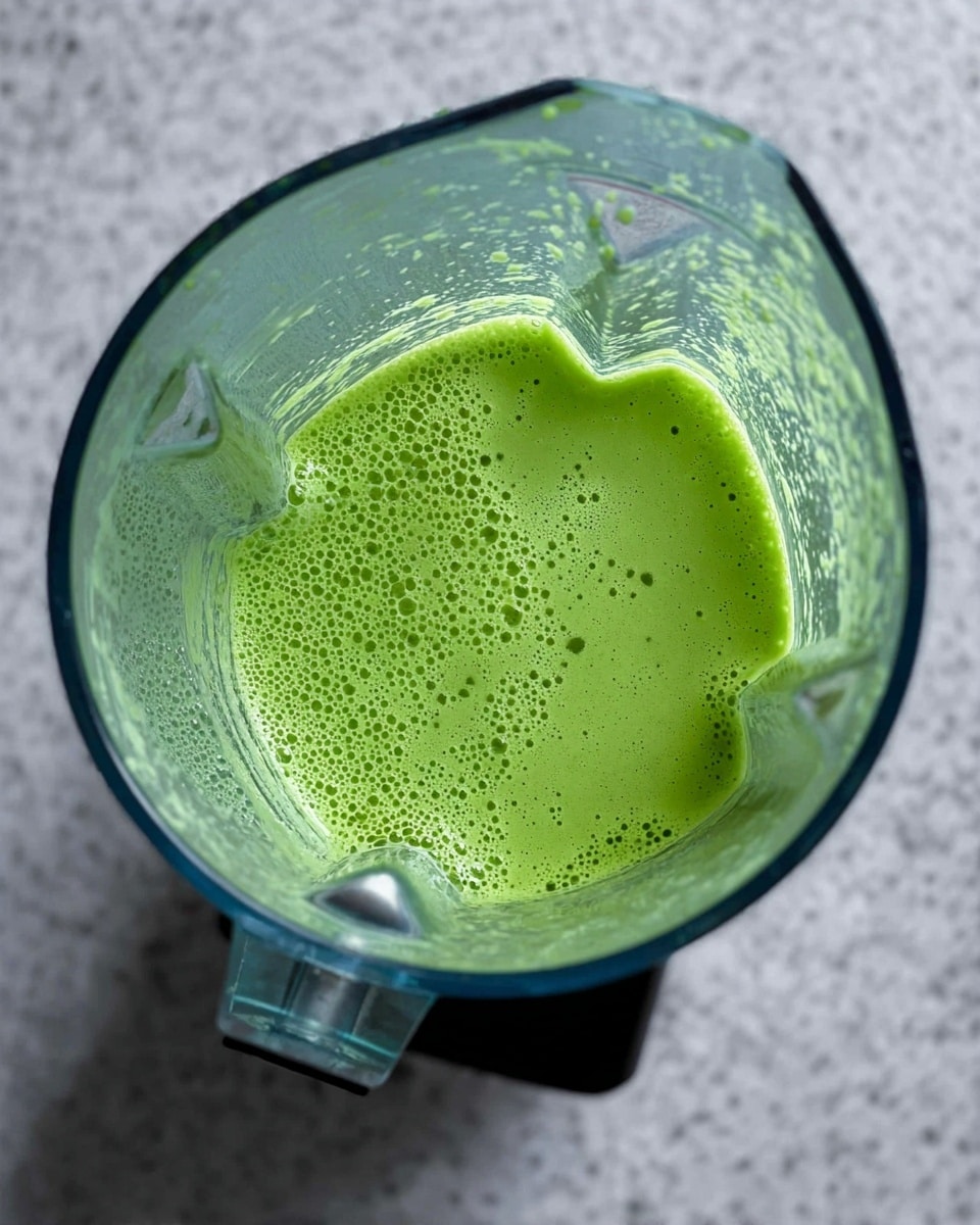 A clear blender container filled with a smooth, bright green liquid topped with small bubbles and foam, showing some texture on the blender sides where the liquid has splashed. The blender is placed on a surface with a white marbled texture. Photo taken with an iphone --ar 4:5 --v 7