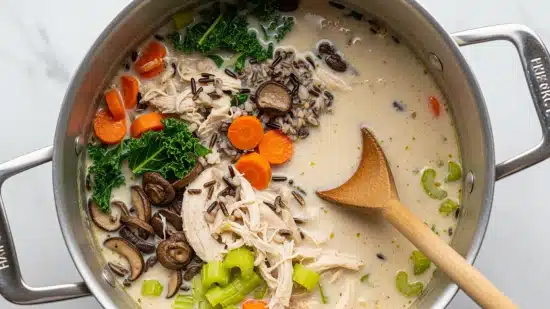 Creamy Wild Rice Chicken Soup with Roasted Mushrooms Recipe 5 A large metal pot filled with creamy white soup showing multiple layers: floating orange carrot chunks, green leafy kale pieces, light beige shredded chicken, some dark wild rice grains, and celery pieces. A wooden spoon with traces of soup rests inside the pot on the right side. The pot is set on a white marbled surface. photo taken with an iphone --ar 4:5 --v 7