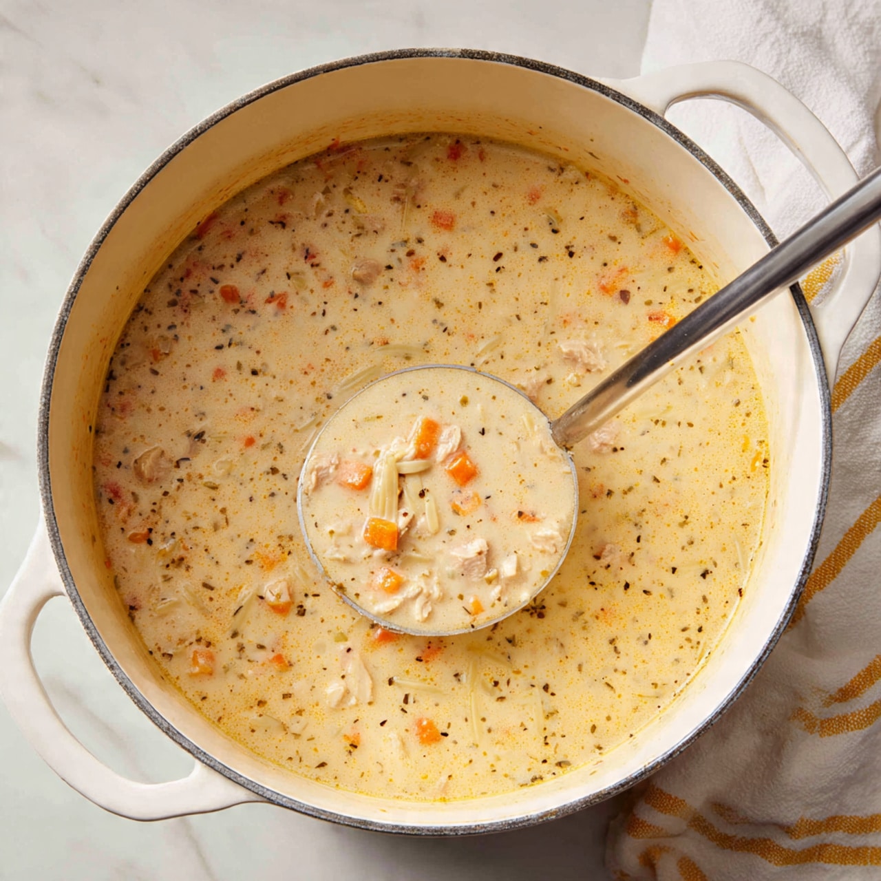 A large white pot filled with creamy soup that is light beige in color with small bits of orange carrot and light purple meat chunks visible throughout. The soup has thin noodle-like bits mixed evenly in the creamy base. A metal ladle is lifted inside the pot showing a close-up of the soup with chunks of carrot and meat surrounded by the thick cream. The pot is set on a white marbled surface with part of a white cloth with yellow stripes visible in the top right corner, photo taken with an iphone --ar 4:5 --v 7