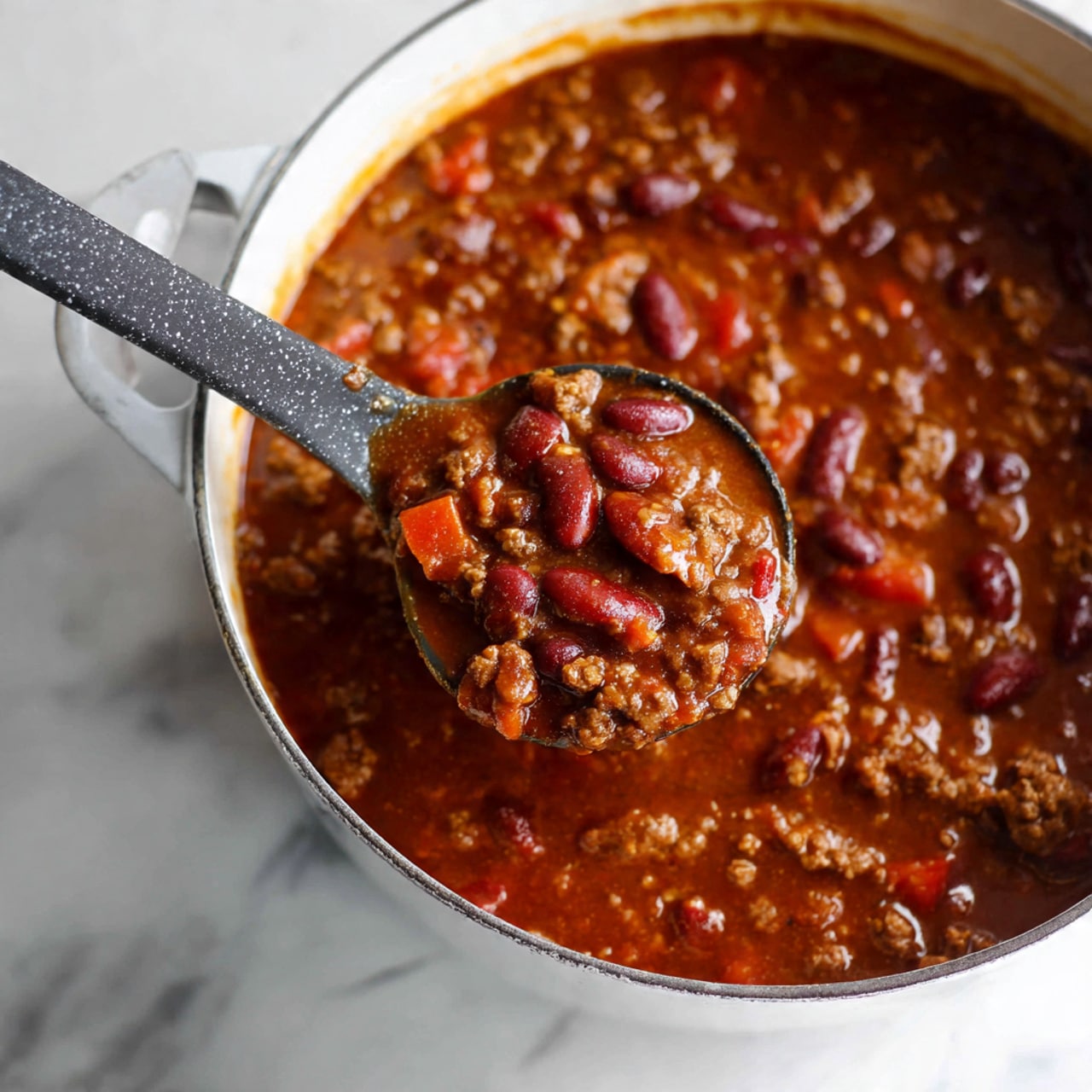 A close-up view of a white pot filled with thick chili, showing a ladle scooping a portion of the chili. The chili has rich red and brown colors with visible kidney beans, small chunks of tomatoes, and ground meat mixed in a thick, glossy sauce. The ladle is black with a gray speckled pattern, dipping into the rich, hearty chili. The background is a white marbled surface. photo taken with an iphone --ar 4:5 --v 7
