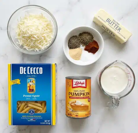 The image shows six cooking items arranged on a white marbled texture. At the top left, there is a clear glass bowl filled with finely shredded white cheese. Next to it on the right, a small white bowl holds five different spices: black pepper, Italian herbs, a reddish-brown spice, garlic powder, and salt, all carefully separated but together in one bowl. To the far right above, there is a wrapped stick of salted butter in cream and yellow packaging. Below these on the left side, a bright blue box of De Cecco penne rigate pasta with yellow edges stands upright, showing some uncooked penne through a clear window at the bottom. Next to it on the right, a can of Libby's 100% pure pumpkin with orange and yellow colors and an image of pumpkin pie on the label is placed. Lastly, in the bottom right corner, a clear glass measuring cup filled with white cream stands with the red measuring scale visible on the side. photo taken with an iphone --ar 4:5 --v 7