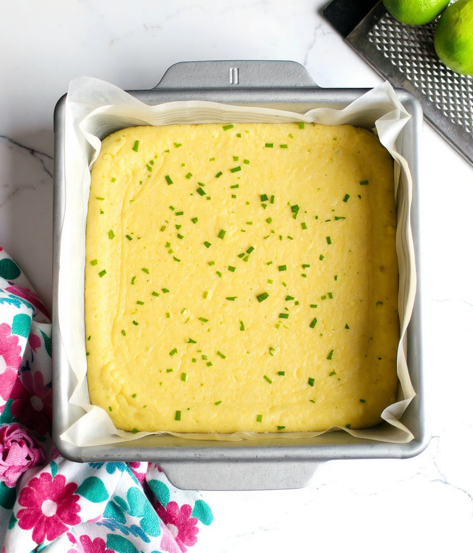 A square metal baking pan lined with white parchment paper holds a single thick layer of smooth, yellow batter with small green zest pieces scattered evenly across the top. The pan rests on a white marbled surface, with a colorful floral cloth underneath its bottom left corner. To the top right beside the pan, there is a metal grater and a dark green lime partially visible, creating a simple kitchen scene. Photo taken with an iphone --ar 4:5 --v 7