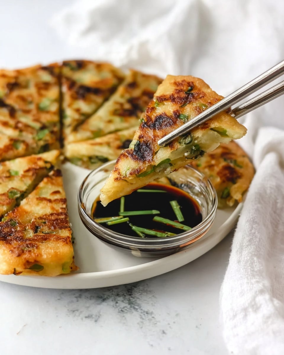 A round, golden-brown pancake sliced into triangular pieces sits on a white plate with one piece held by metal chopsticks above a clear glass bowl filled with dark soy sauce mixed with green onion slices. The pancake has visible green onion pieces inside and a slightly crispy texture with charred spots on the surface. The background is a white marbled texture, and a white cloth napkin is softly visible in the background. photo taken with an iphone --ar 4:5 --v 7