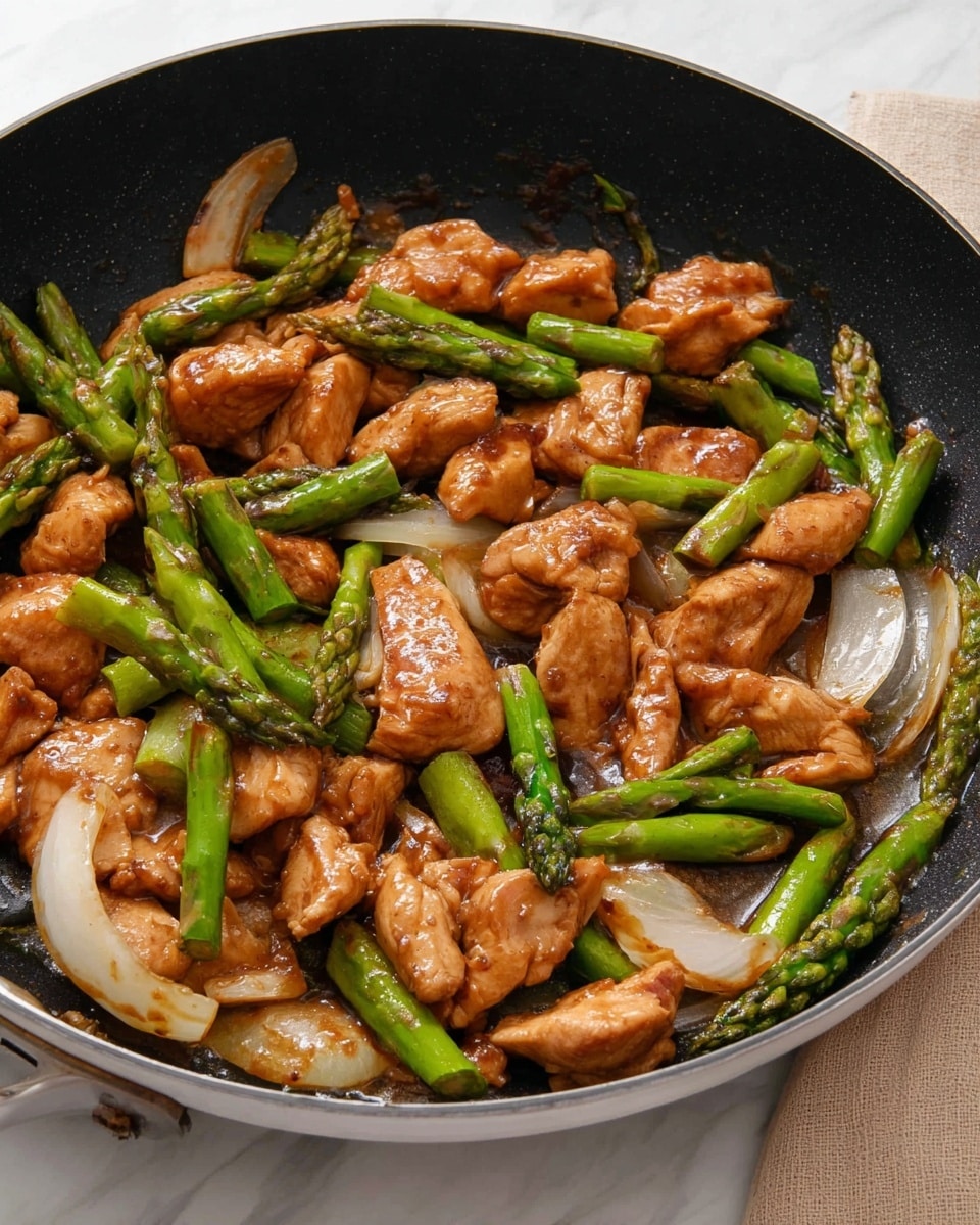 The image shows a close-up of a black pan filled with cooked pieces of light brown chicken mixed with bright green asparagus tips and thick slices of white onion. The chicken pieces have a glossy, slightly shiny texture from a brown sauce covering them, and the asparagus appears crisp and fresh. The onion slices are scattered among the chicken and asparagus, adding a contrasting pale color. The pan sits on a white marbled surface with a beige cloth partially visible in the background. photo taken with an iphone --ar 4:5 --v 7