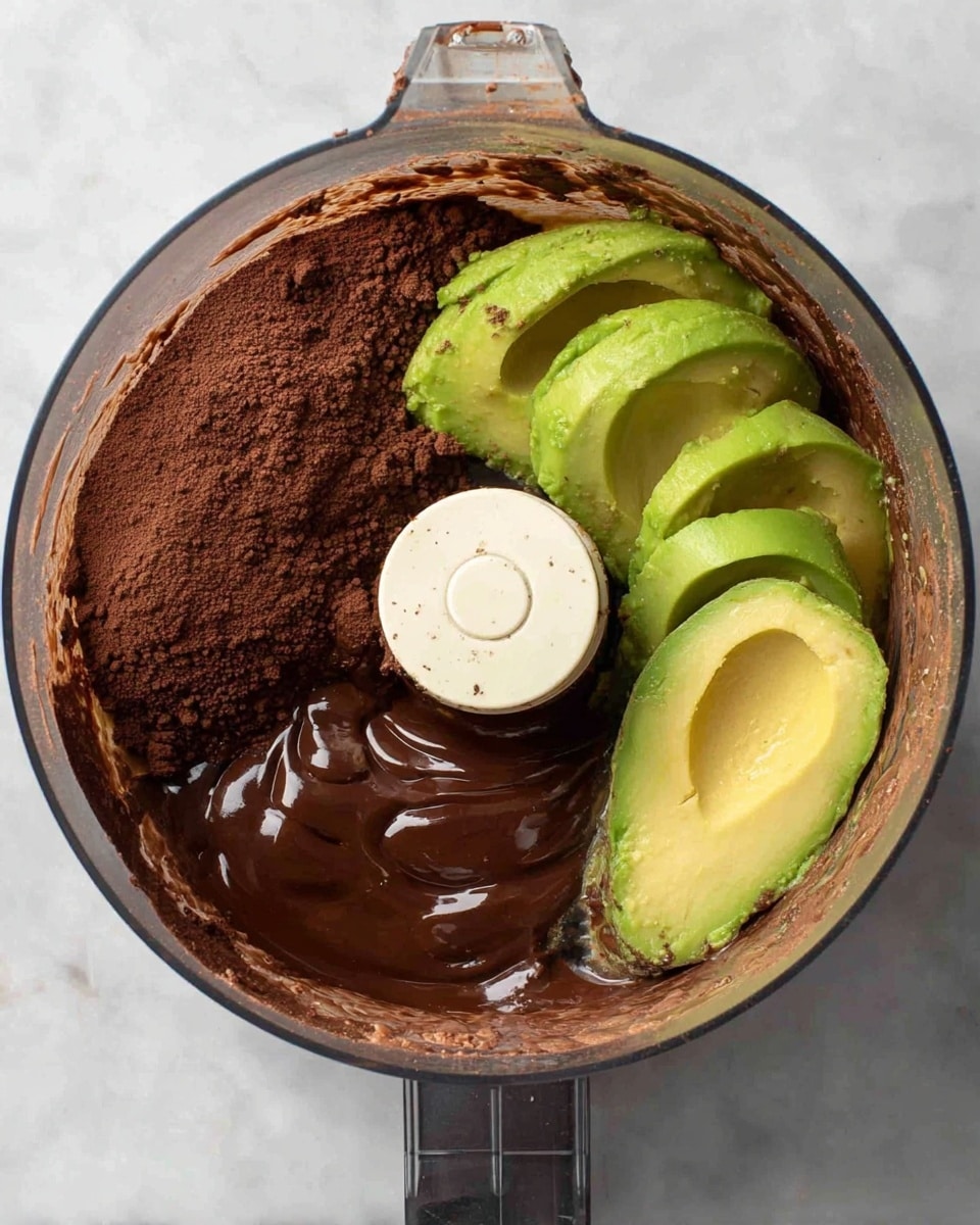 A clear glass food processor bowl contains three main layers of ingredients arranged separately: dark brown cocoa powder spread across the bottom, glossy melted chocolate thickly poured over part of the cocoa, and three bright green halved avocados placed on the opposite side. The food processor blade is white and centered, contrasting with the darker ingredients. The background is a white marbled surface. Photo taken with an iphone --ar 4:5 --v 7