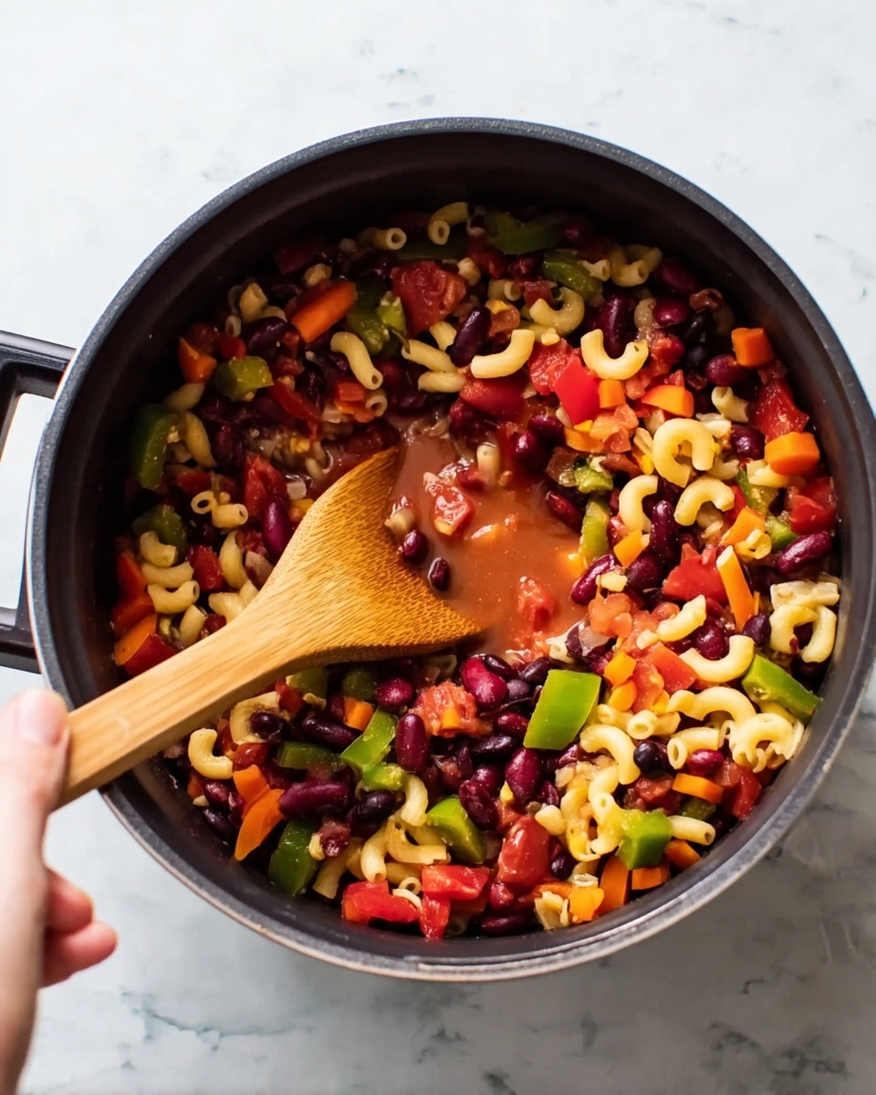 A black pot filled with a colorful mix of food ingredients including kidney beans, black beans, elbow macaroni pasta, diced red tomatoes, carrot slices, and green pepper pieces. The mixture looks slightly wet with some sauce or juice visible at the center. A woman's hand is holding a light wooden spoon inside the pot, stirring the colorful contents. The pot is placed on a white marbled surface photo taken with an iphone --ar 4:5 --v 7
