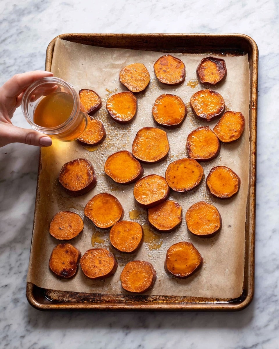 A baking tray lined with parchment paper holds about 18 round slices of roasted orange sweet potatoes, showing a slightly browned and crispy surface with juicy textures shining under light. The slices are spread out evenly in a few scattered rows, with some darker spots indicating caramelization. A woman's hand is pouring a light amber syrup over one of the sweet potato slices from a small glass container. The tray is placed on a white marbled surface that adds a clean and simple backdrop to the warm colors of the food photo taken with an iphone --ar 4:5 --v 7