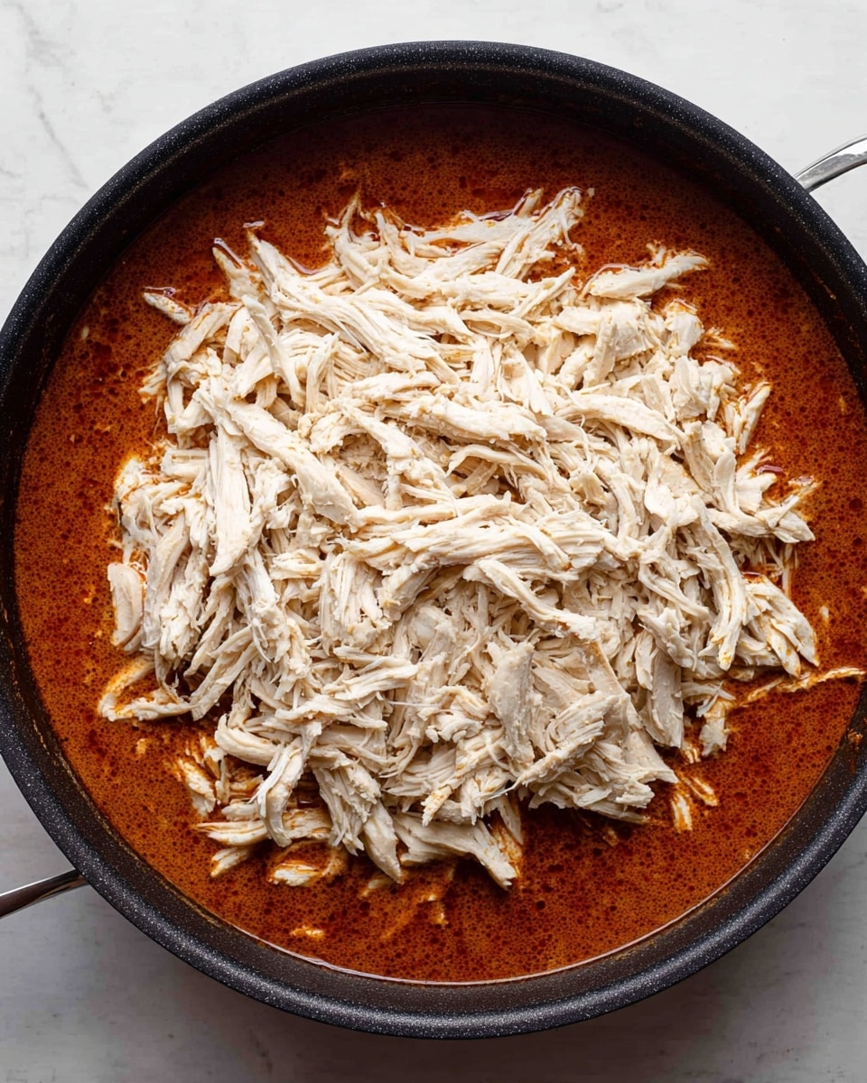 A black pan filled with two layers: the bottom layer is a smooth, rich reddish-brown sauce with a slightly oily surface, and the top layer is a large mound of pale, shredded chicken spread evenly in the center of the sauce, covering most of the pan. The pan sits on a white marbled surface. photo taken with an iphone --ar 4:5 --v 7