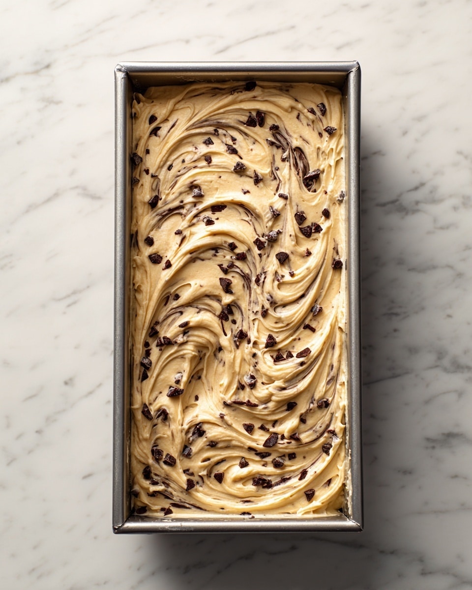 A gray metal baking tray filled with one even layer of light beige batter mixed with small dark chocolate chips throughout, the batter having a smooth and slightly shiny texture with visible swirls and folds on top, placed on a white marbled surface photo taken with an iphone --ar 4:5 --v 7