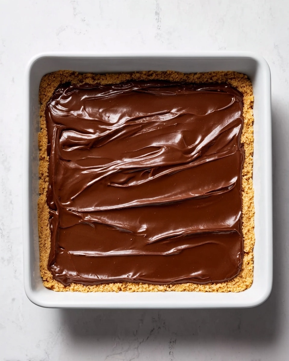 The image shows a white rectangular baking dish filled with a dessert that has two clear layers. The bottom layer is a light golden brown crumbly crust that lines the entire dish evenly and slightly uneven at the edges. The top layer is a thick, glossy, smooth dark chocolate spread covering the crust fully, with some soft wave-like swirls and gentle ridges visible on the surface. The dish is placed on a white marbled textured surface. photo taken with an iphone --ar 4:5 --v 7