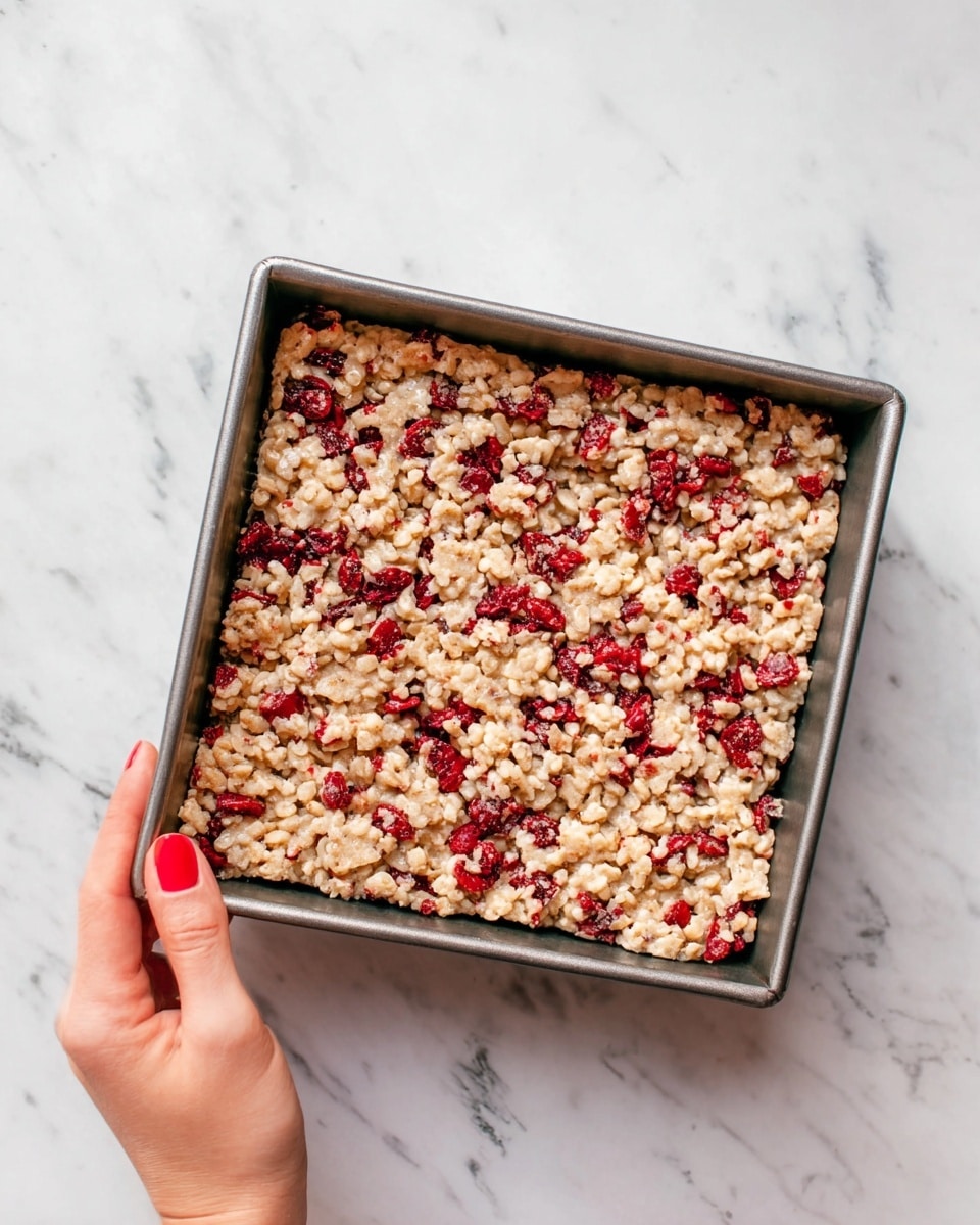 A square metal baking pan filled with one thick layer of light beige cereal bars mixed with bright red pieces of dried fruit, giving it a slightly bumpy texture. A woman's hand with red nail polish is holding the left side of the pan. The pan is set on a white marbled surface. photo taken with an iphone --ar 4:5 --v 7