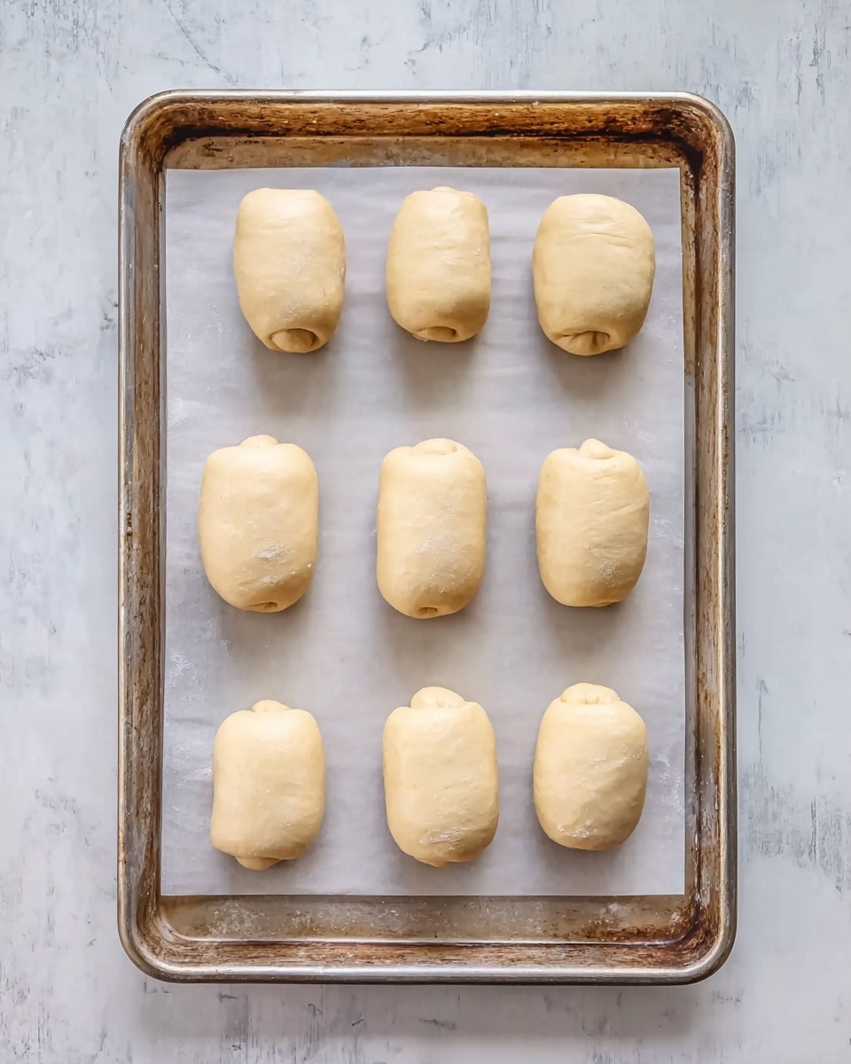 A metal baking tray lined with white parchment paper holds eight small dough rolls arranged in three rows. Each dough roll is light beige and smooth with a slightly rounded shape, some having a small end tucked underneath. The tray rests on a surface with a white marbled texture. photo taken with an iphone --ar 4:5 --v 7