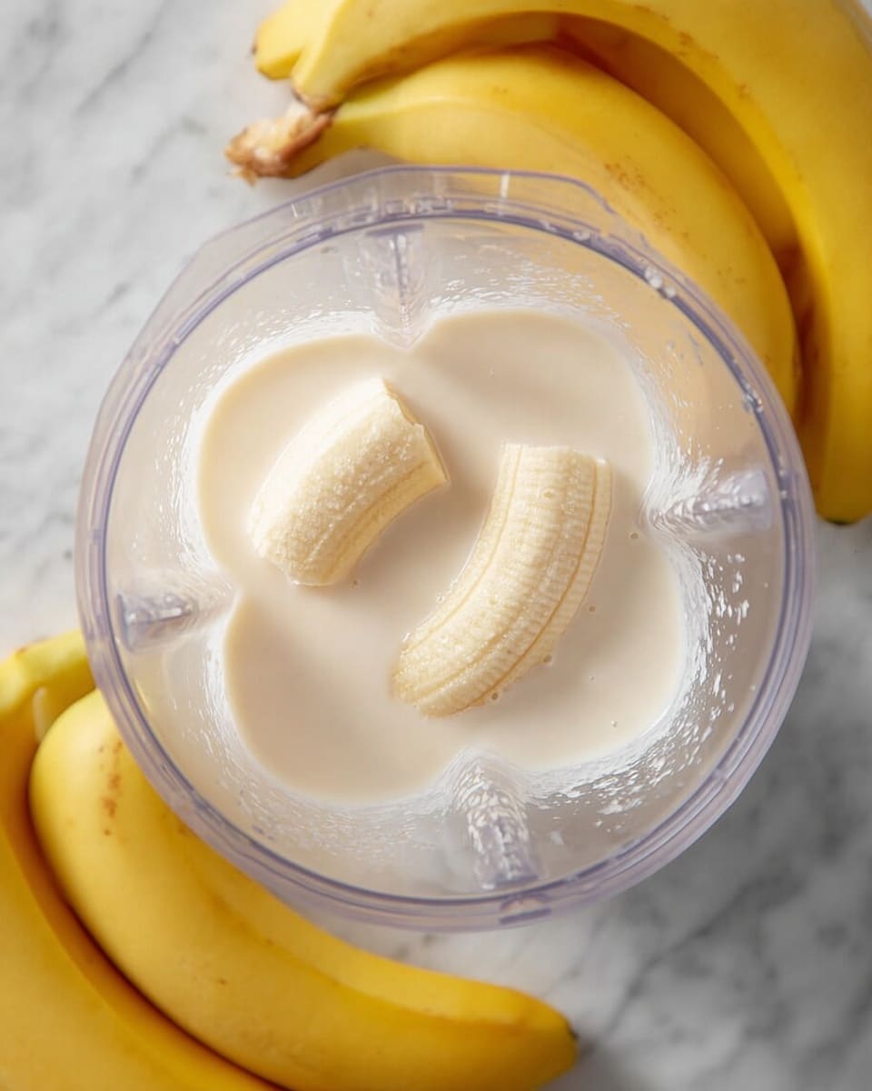 A clear blender container is shown from above with two peeled banana halves that are light yellow and soft in texture lying in the middle of a thick, creamy, off-white liquid which fills about one-third of the container's base. Around the blender, there are several whole yellow bananas resting on a white marbled surface. The image has soft natural light highlighting the smooth texture of the liquid and the bananas, emphasizing freshness. photo taken with an iphone --ar 4:5 --v 7