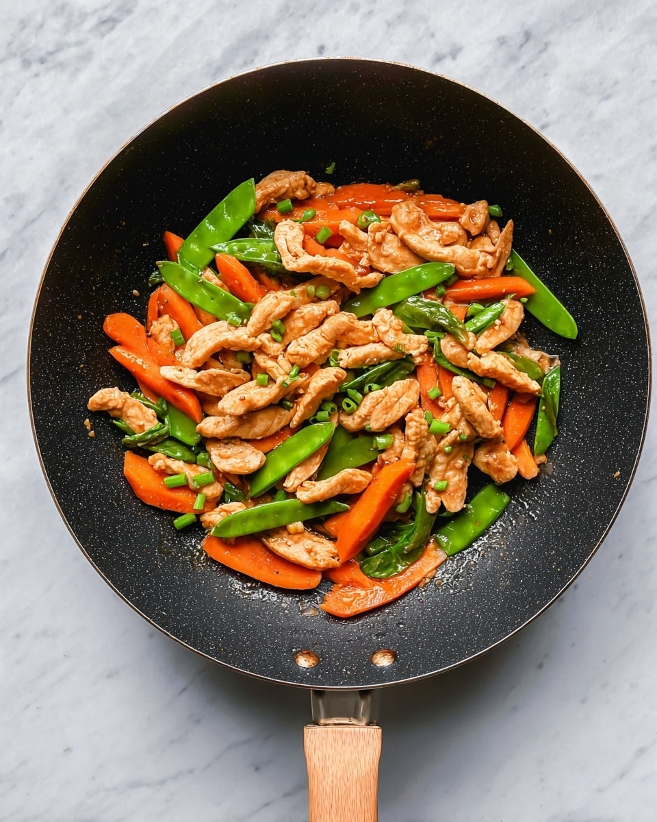 A black frying pan with a wooden handle holds a stir-fry of three layers: the first layer is light brown strips of cooked chicken spread evenly across the pan; the second layer includes bright orange carrot slices and green snap peas mixed throughout; the third layer is small chopped green onions sprinkled on top, adding a fresh touch. The pan sits on a white marbled surface. photo taken with an iphone --ar 4:5 --v 7