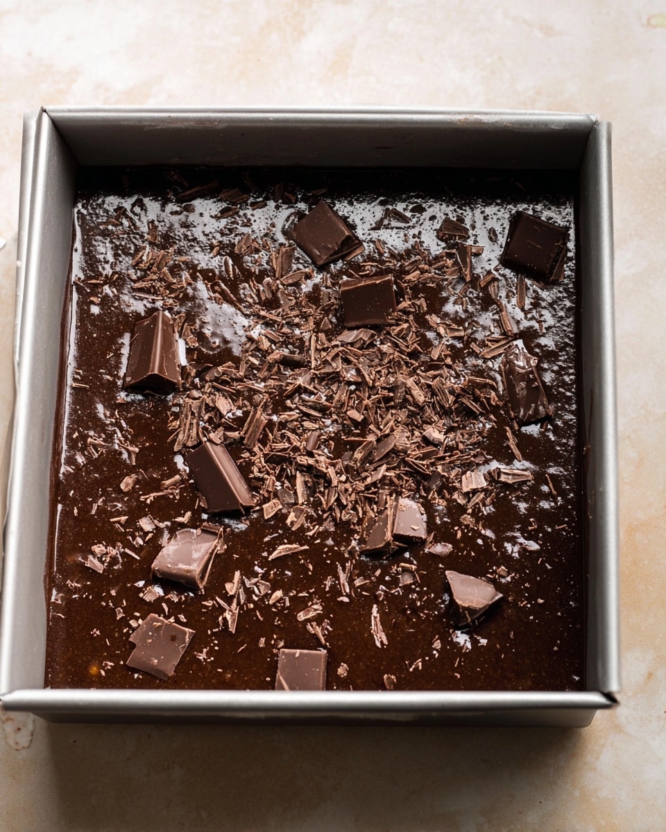 A square metal baking pan filled with a dark brown chocolate batter that has a shiny, wet texture. The top layer shows unevenly scattered pieces of chopped chocolate, some larger and some finely grated, resting on the smooth surface of the batter. The pan sits on a white marbled surface with a soft, light tone. The photo taken with an iphone --ar 4:5 --v 7
