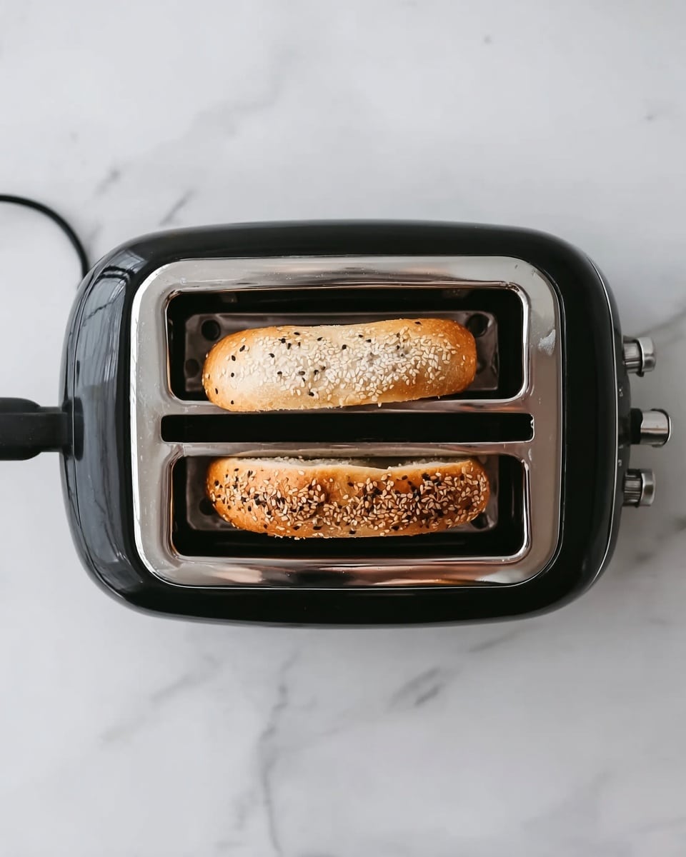 The image shows a black toaster with a shiny silver top, placed on a white marbled surface. Inside the two slots of the toaster, two halves of a bagel are visible. The top half is plain with a light golden brown crust, while the bottom half is covered with sesame and other seeds, toasted to a warm golden color. The toaster's power cord extends from the left side. The overall scene is bright and clear, highlighting the texture of the bagels and the sleek design of the toaster. photo taken with an iphone --ar 4:5 --v 7