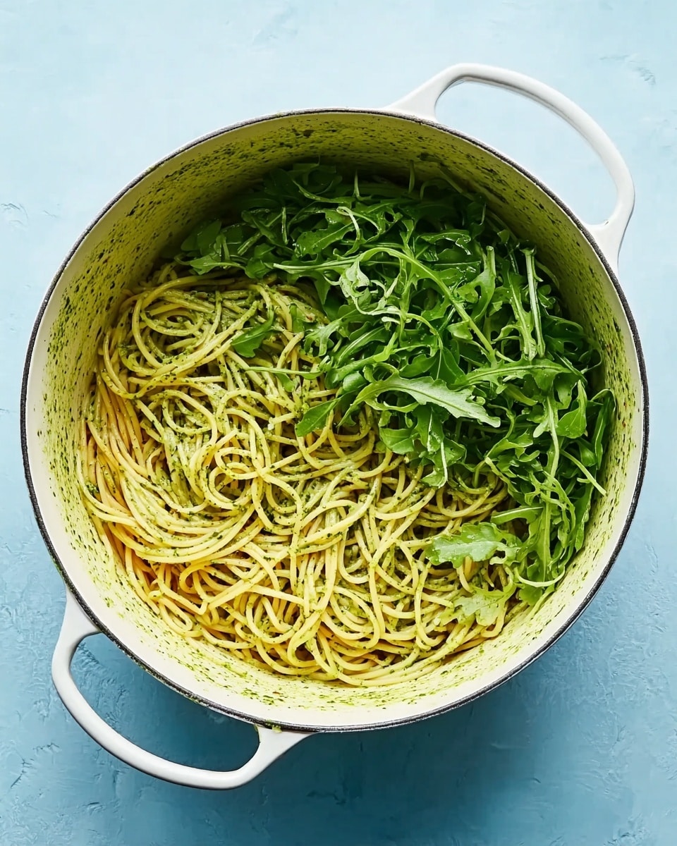 A large white pot sits on a white marbled surface, filled halfway with thin yellow spaghetti noodles mixed with a green herb sauce, and the other half is covered with fresh bright green arugula leaves. The pot has two handles on each side, and the inside edges show some green sauce stains. The background is a soft blue color, which contrasts with the colors inside the pot. photo taken with an iphone --ar 4:5 --v 7