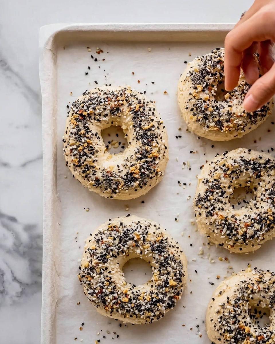 The image shows four pale, round bagels with a hole in the center, placed on a white baking sheet on a white marbled surface. Each bagel has a rough texture and is covered with a mix of black and white sesame seeds and small bits of seasoning. A woman's hand is seen on the top right side, sprinkling more seeds onto one of the bagels. The scene captures a close view, highlighting the uneven spread of seeds and the soft dough texture before baking. Photo taken with an iphone --ar 4:5 --v 7