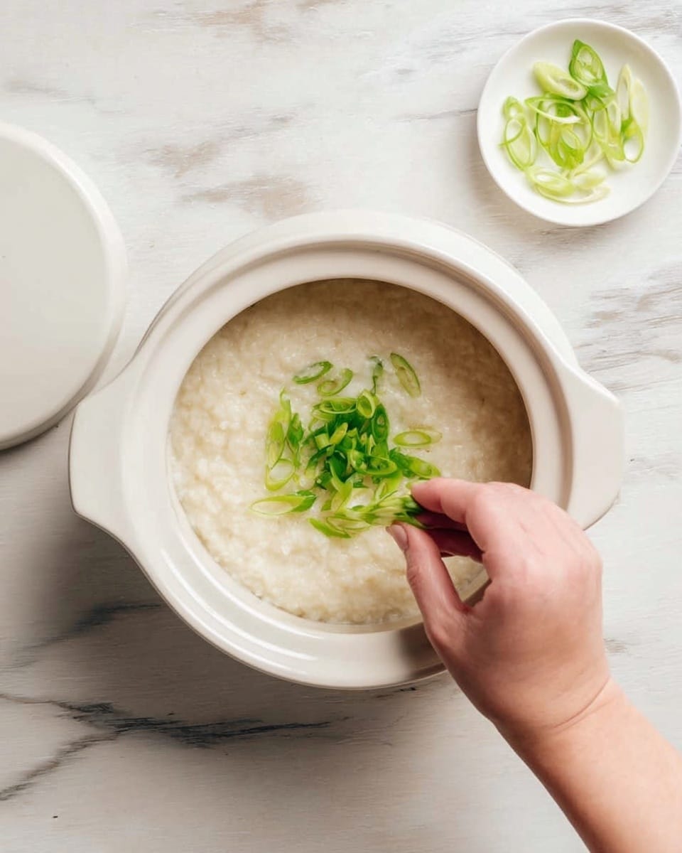A white ceramic pot holds a creamy, light beige rice porridge with a thick, smooth texture. On top, bright green sliced scallions are being sprinkled by a woman's hand, adding a fresh contrast to the pale porridge. A small white dish with more scallions sits nearby on a white marbled surface. The overall scene is simple and clean with soft natural light. photo taken with an iphone --ar 4:5 --v 7