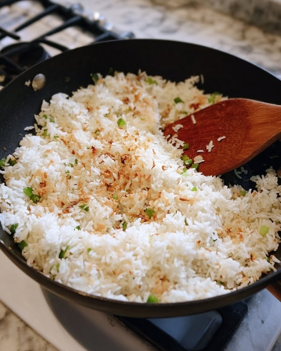 A close-up photo of a black frying pan filled with white rice, lightly toasted brown bits, and small pieces of green onion mixed in. A wooden spoon is stirring the rice on the right side of the pan. The pan sits on a stove with a white marbled surface visible in the background. photo taken with an iphone --ar 4:5 --v 7