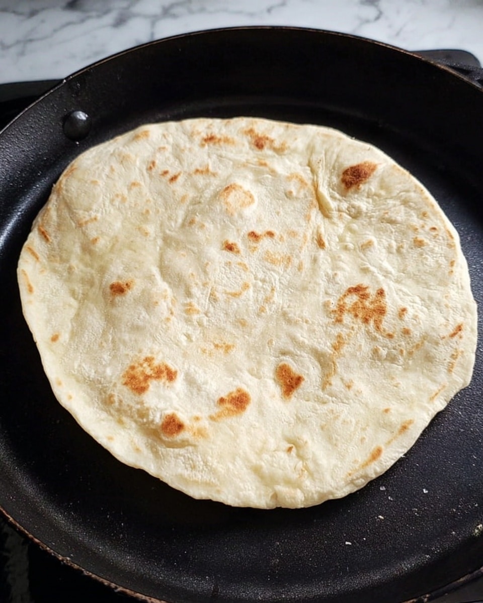 A single round flatbread or tortilla is cooking on a black skillet. The flatbread is light beige with a soft, slightly wrinkled texture and has a few small brown spots scattered on its surface from cooking. The skillet has a matte finish and is placed on a stove. The background surface shown is a white marbled texture. Photo taken with an iphone --ar 4:5 --v 7