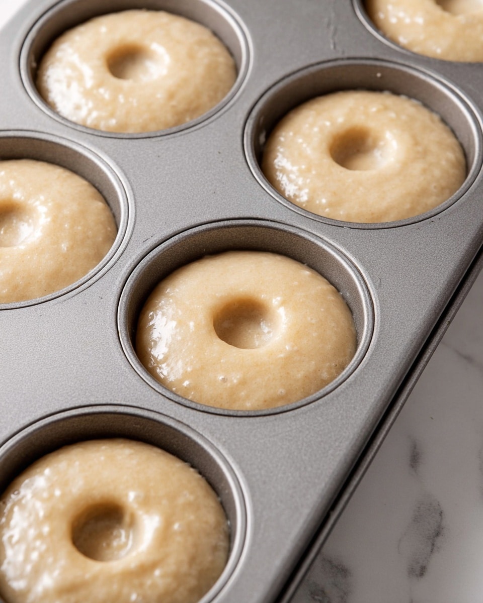 A close-up look at a gray metal donut baking pan filled with light beige batter. Each donut cup holds one layer of smooth, creamy batter with small air bubbles on the surface. The batter fills the cups almost to the top, covering the flat center post in each cup. The pan is placed on a white marbled surface giving a clean and bright background. photo taken with an iphone --ar 4:5 --v 7