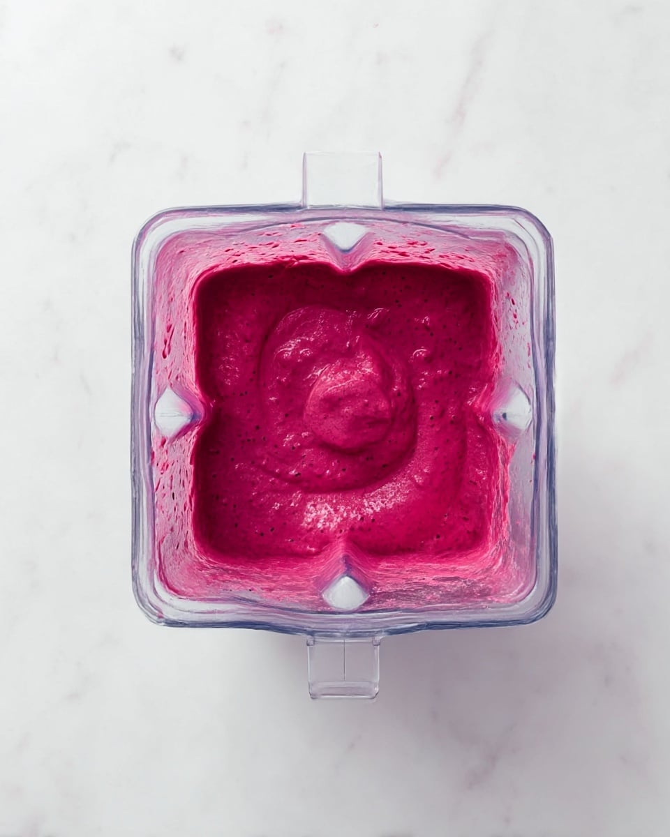 A top-down view of a blender containing a thick, bright magenta mixture swirling around the blade. The blender is clear plastic, revealing the chunky, textured blend inside. The scene is set on a smooth white marbled surface, which contrasts with the rich, bold color of the mixture. No other objects are visible, and the focus is entirely on the vibrant mixture in motion. photo taken with an iphone --ar 4:5 --v 7