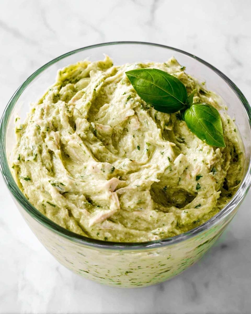 The image shows a clear glass bowl filled with a creamy, textured mix that has a light green color with bits of white mixed throughout. The mix appears thick and slightly chunky, with visible small pieces and a swirled pattern on top. A fresh green basil leaf rests on the surface, adding a bright splash of color. The bowl sits on a white marbled surface. photo taken with an iphone --ar 4:5 --v 7