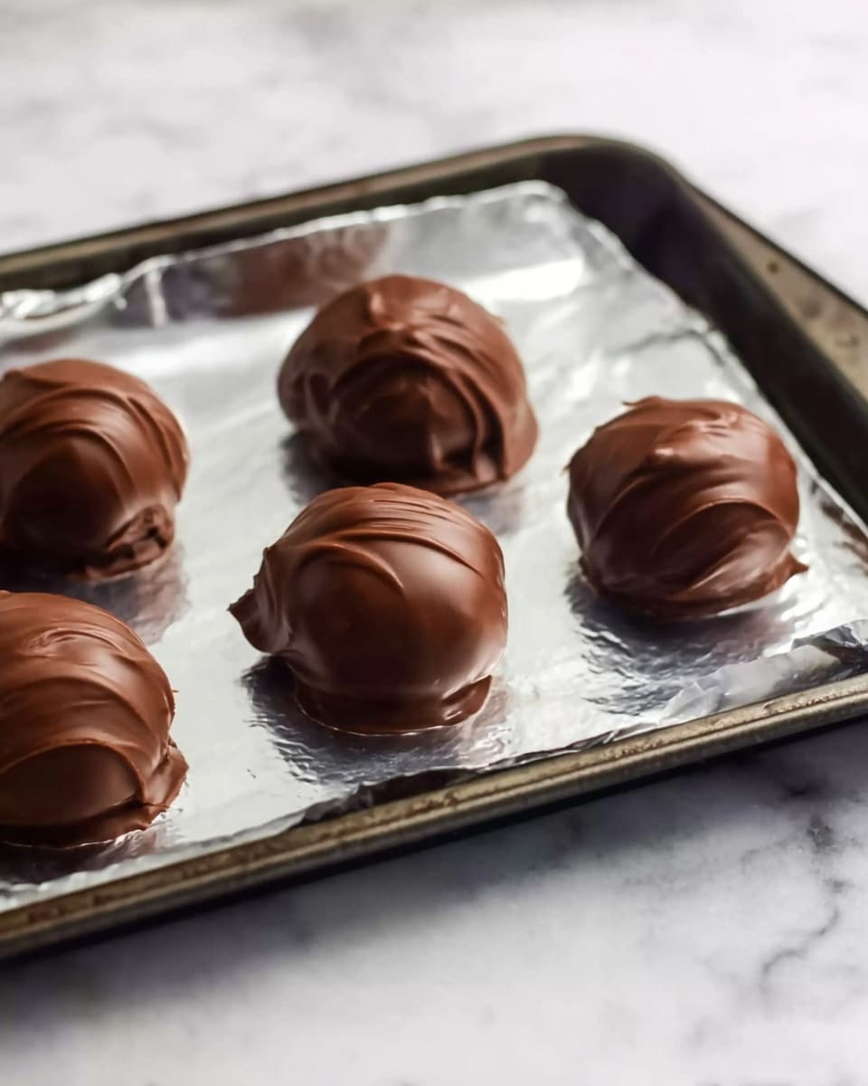 The image shows six round chocolate balls placed on a baking tray lined with silver foil. Each ball is covered with smooth, shiny dark brown chocolate coating that has slight swirls and ripples on the surface. The balls are evenly spaced in two rows with three in the first row and three in the second row. The tray has a dark metal edge and sits on a white marbled surface. The lighting creates soft reflections on the chocolate, making it look fresh and glossy. Photo taken with an iphone --ar 4:5 --v 7
